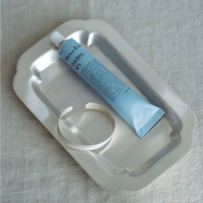 A light blue tube of hand balm rests inside a white scalloped metal dish on a textured beige linen background.