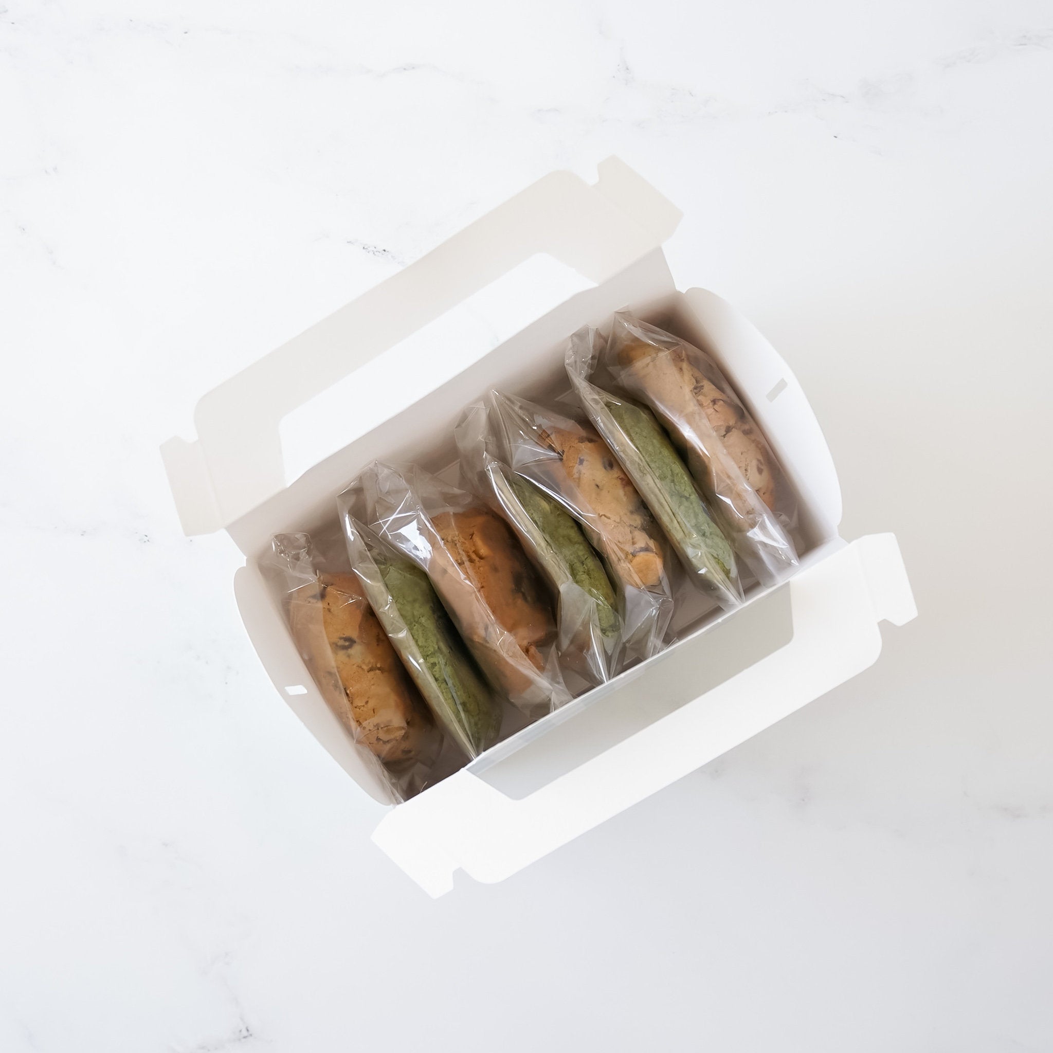 A white cardboard box with a handle, containing seven cookies. The cookies are individually wrapped in clear plastic. Three cookies are green and four cookies are brown.