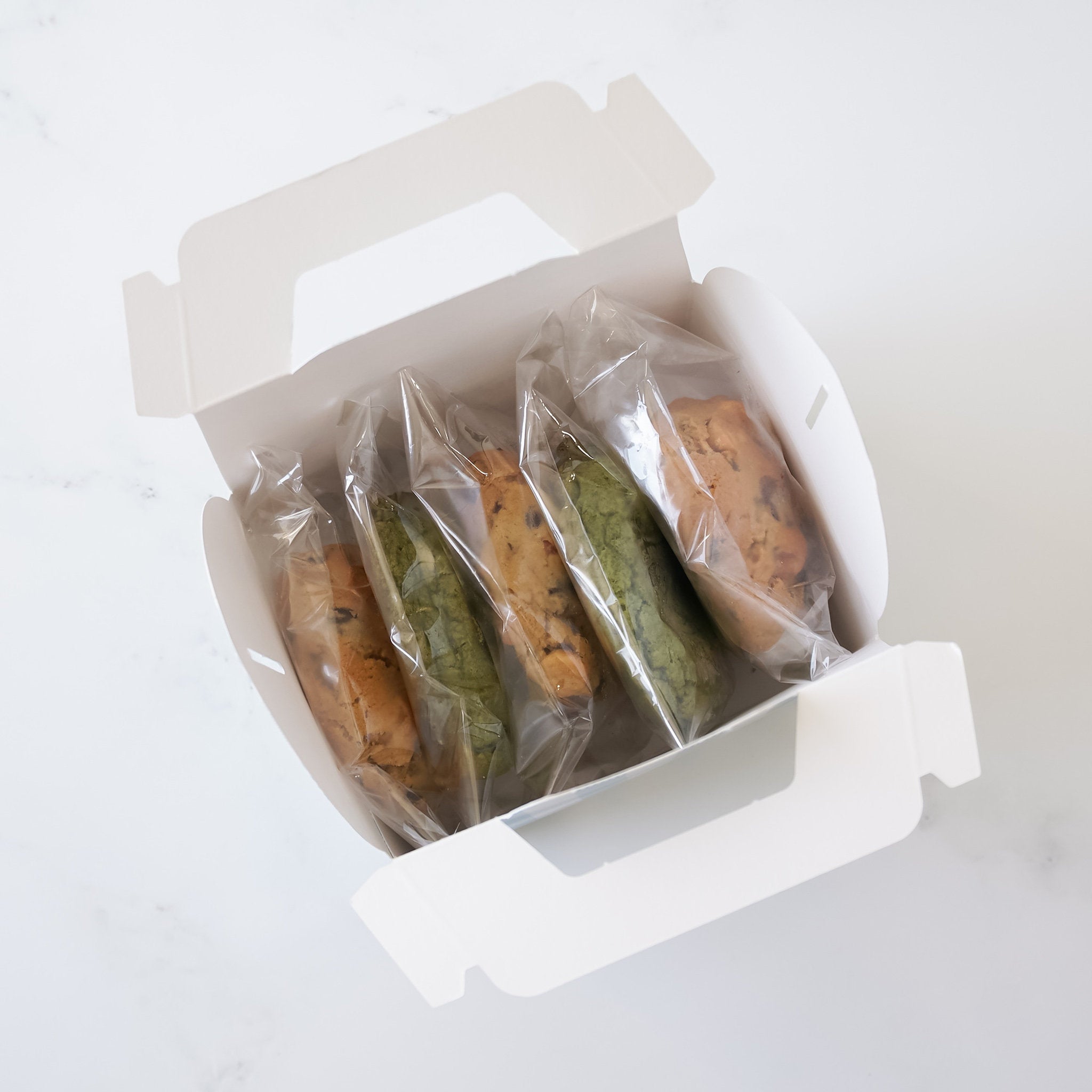 A white cardboard box with a handle, containing five cookies. The cookies are individually wrapped in clear plastic. Two cookies are green and three cookies are brown.