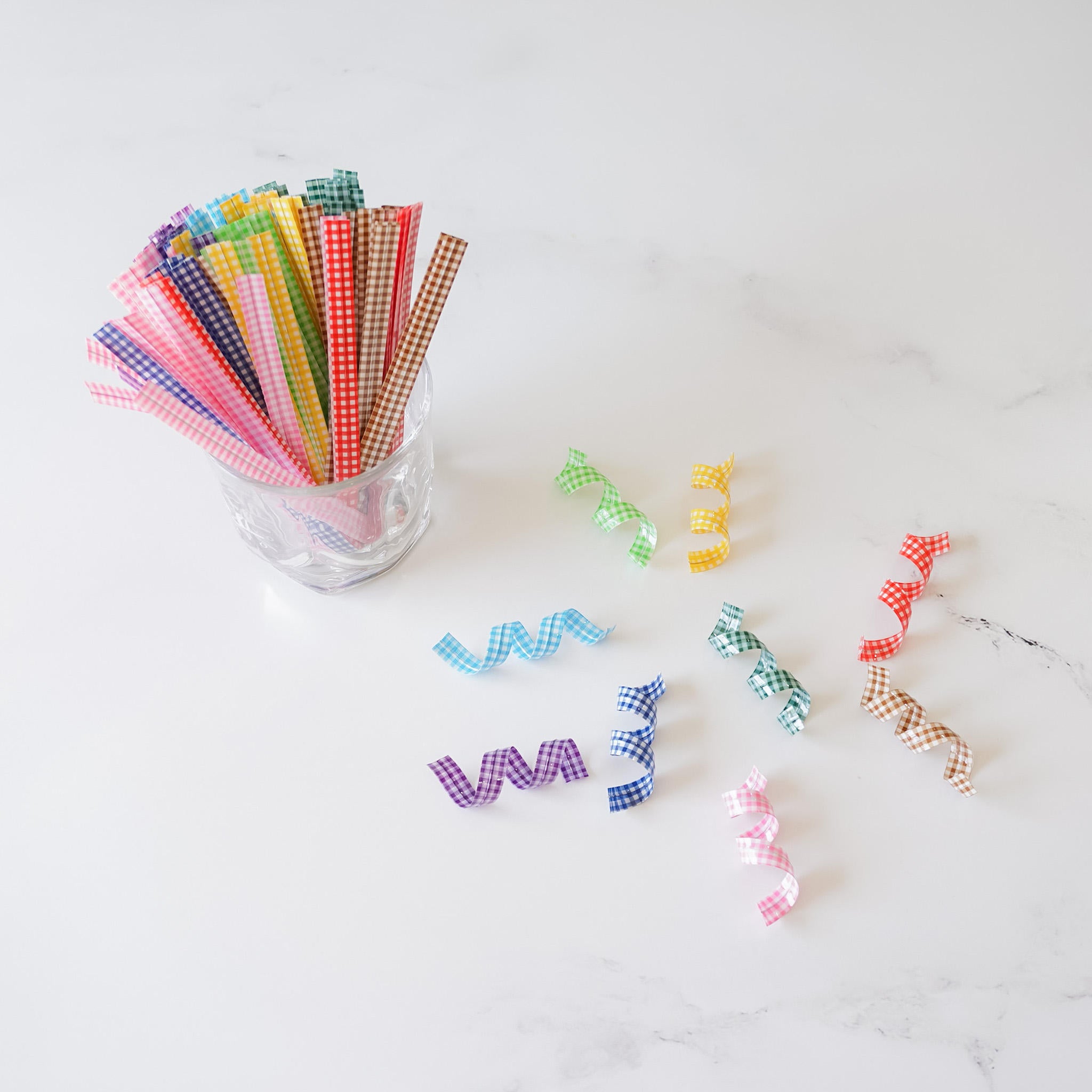 A collection of colorful gingham check twist ties arranged on a white surface, with a glass container filled with ties in the background.