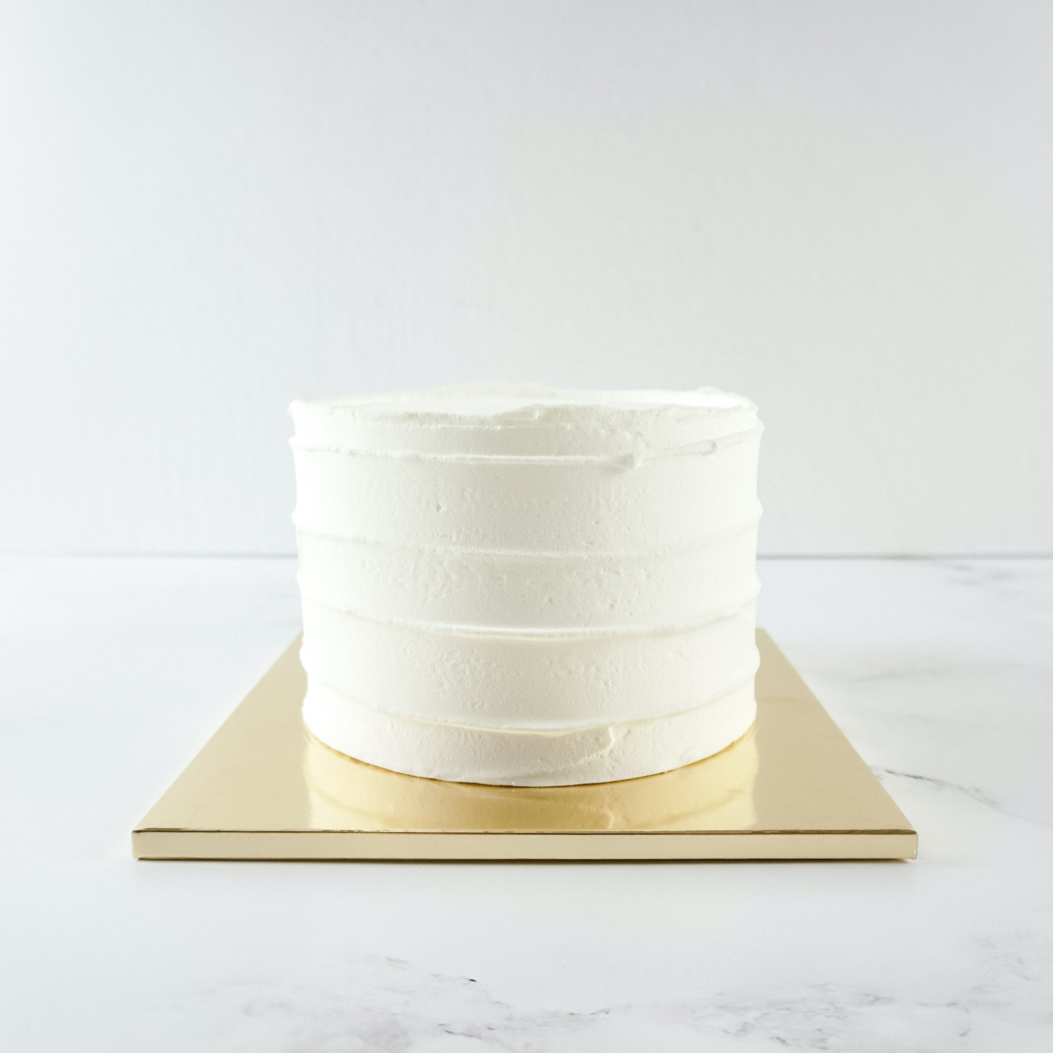 A white cake with a smooth, slightly textured frosting. The cake is sitting on a gold cake board.