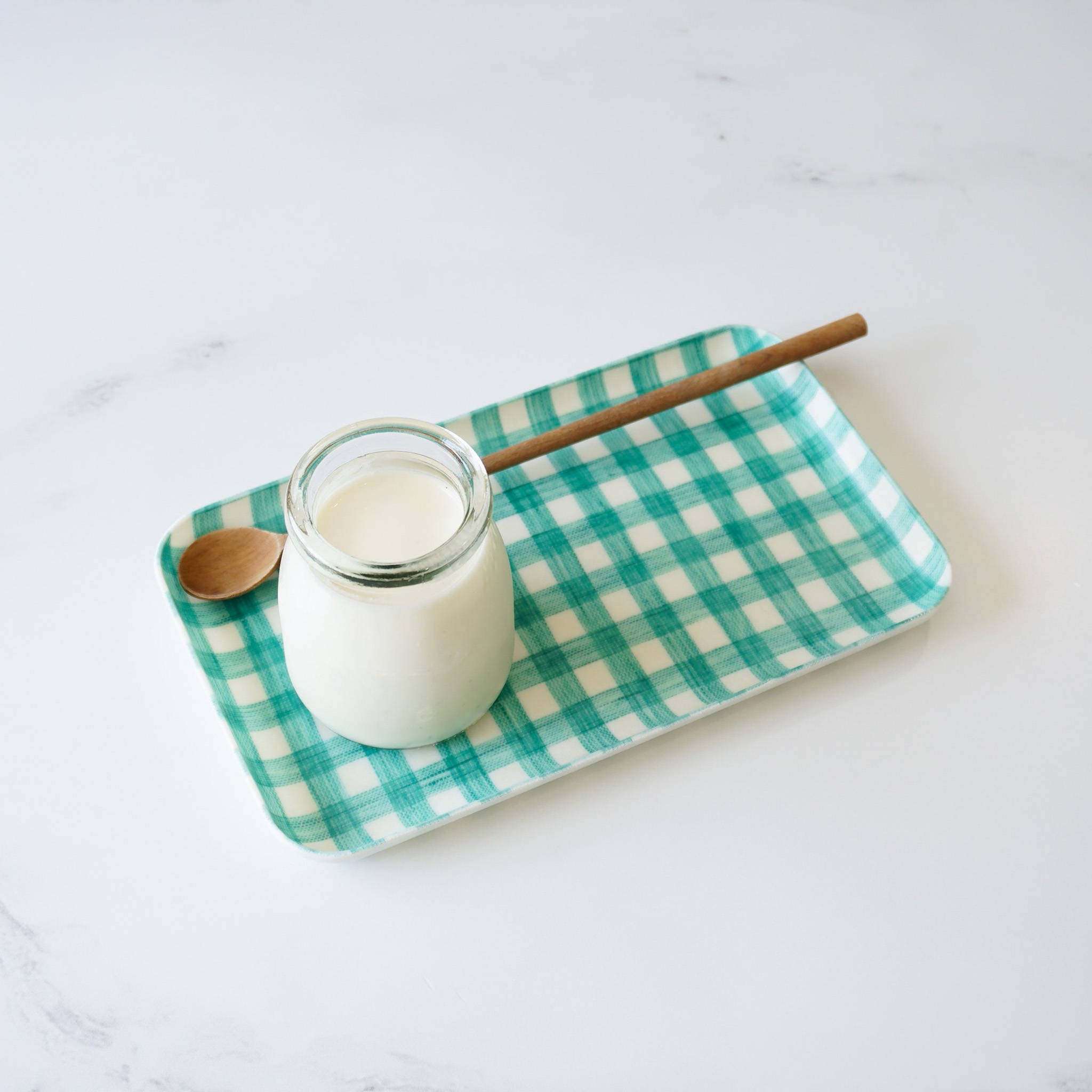 A green check linen coated tray with a glass jar and a wooden spoon on it, placed on a white surface.