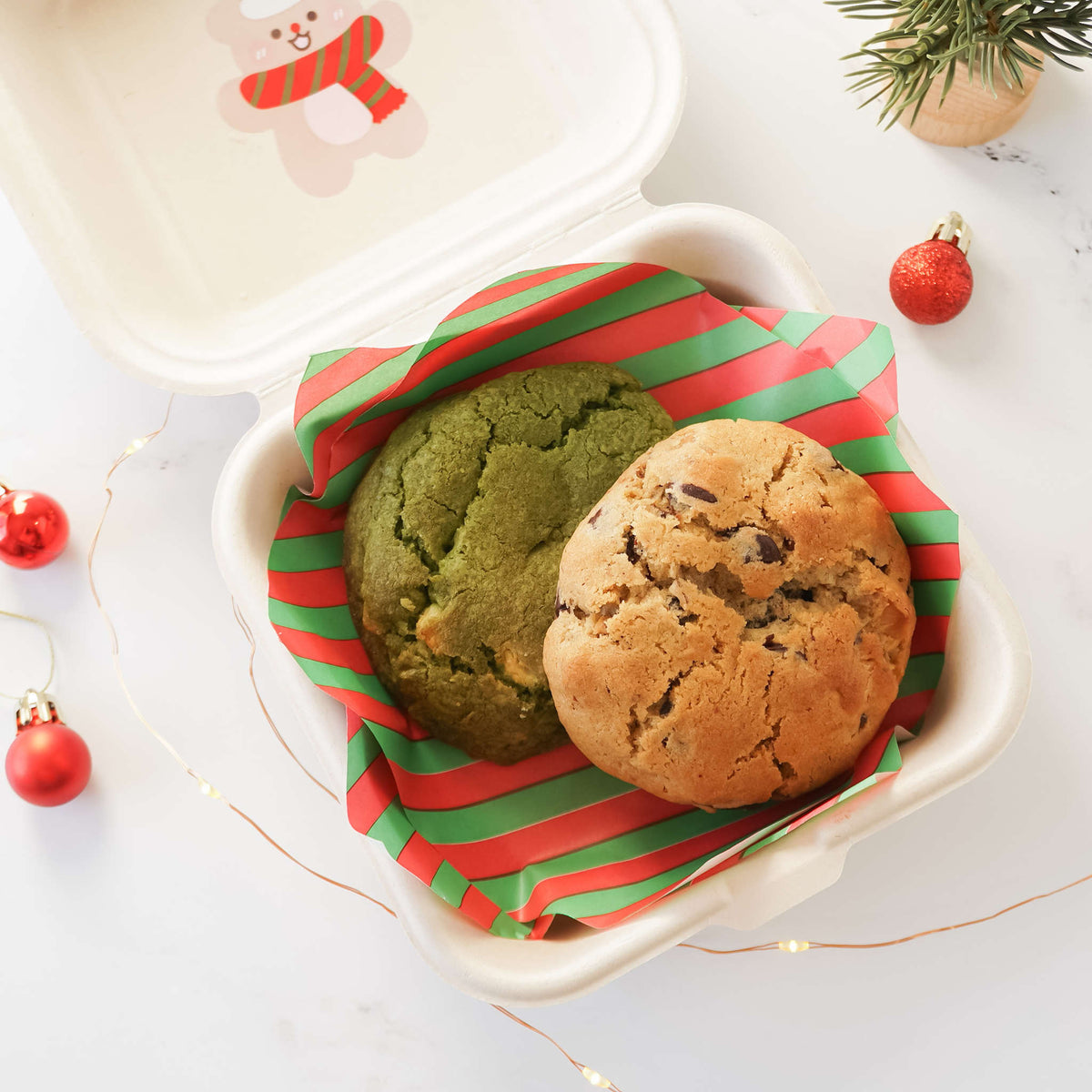 A deli paper with red and green stripes, featuring two cookies on top, placed inside a white container with a Christmas snowman design. Holiday decorations are visible around the setting.