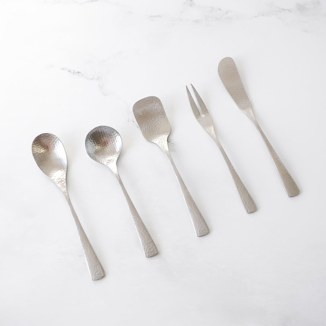 A set of five hammered stainless steel dessert utensils. The set includes a spoon, a round spoon, a square-ended spoon, a fork, and a butter knife. The utensils are arranged on a white surface.