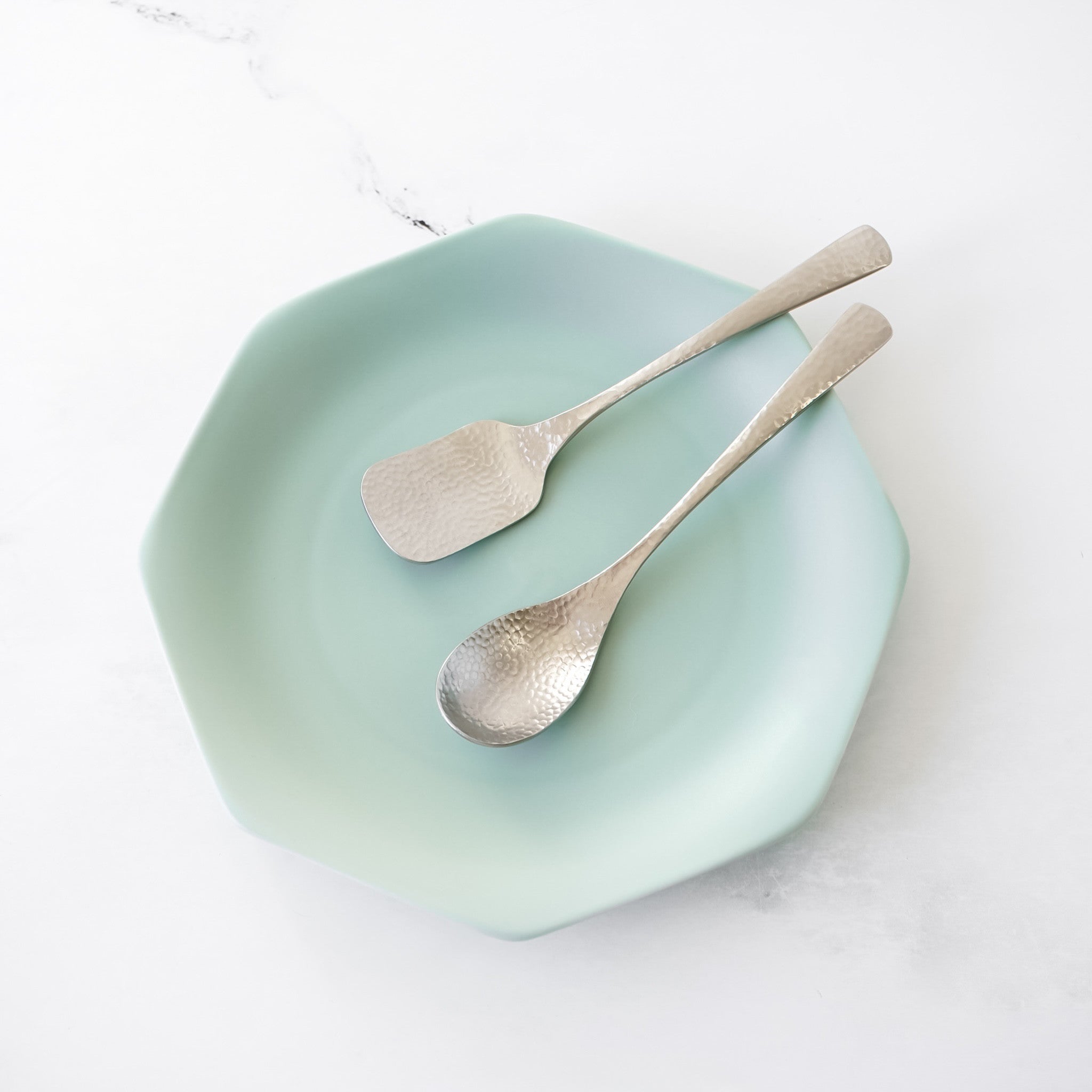 A light green, octagonal plate with two silver dessert utensils. One utensil is a teaspoon, and the other is a square-shaped spoon. The utensils have a hammered texture. The plate and utensils are on a white surface.