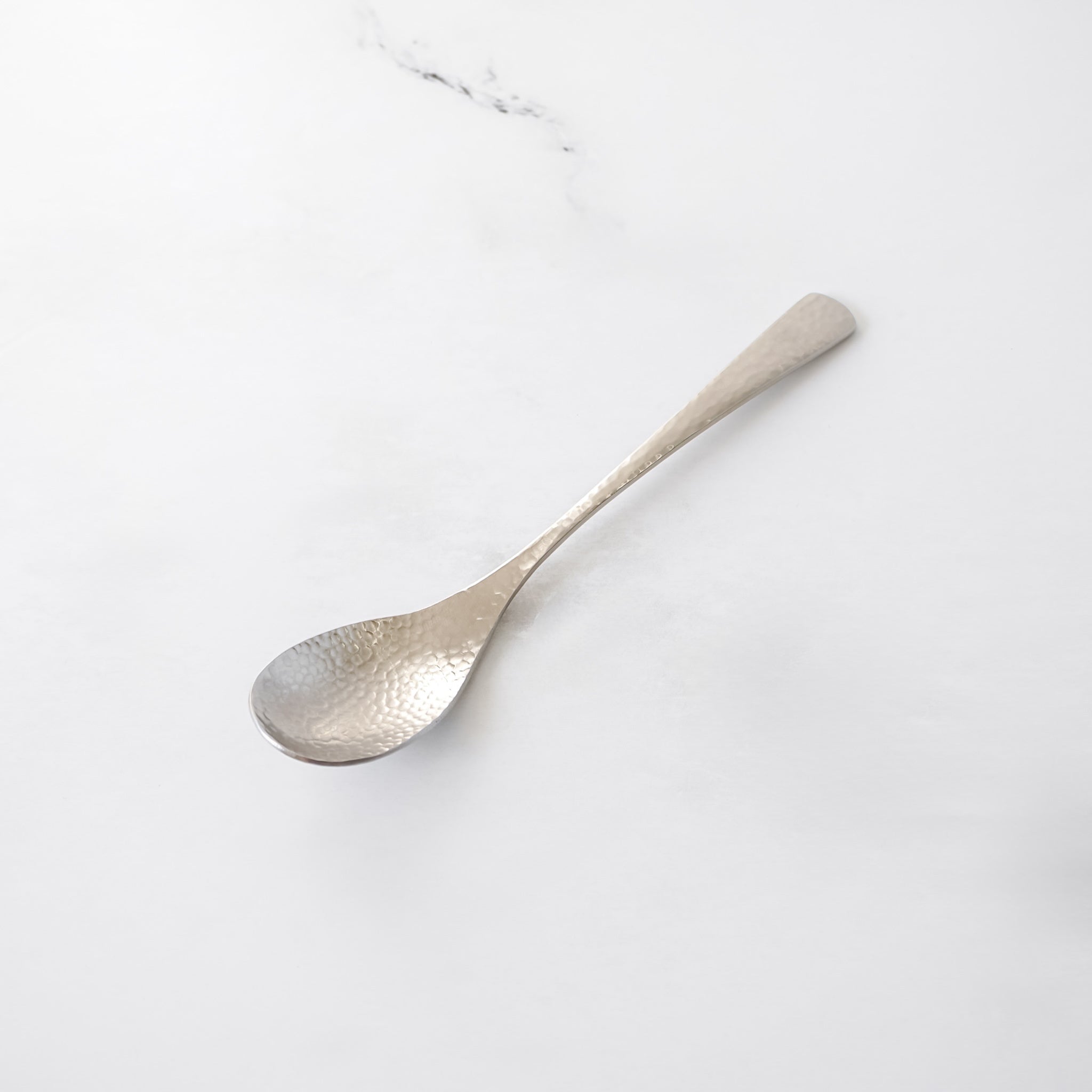 A silver spoon with a hammered texture. The spoon has a long, slender handle and is set against a white background. 