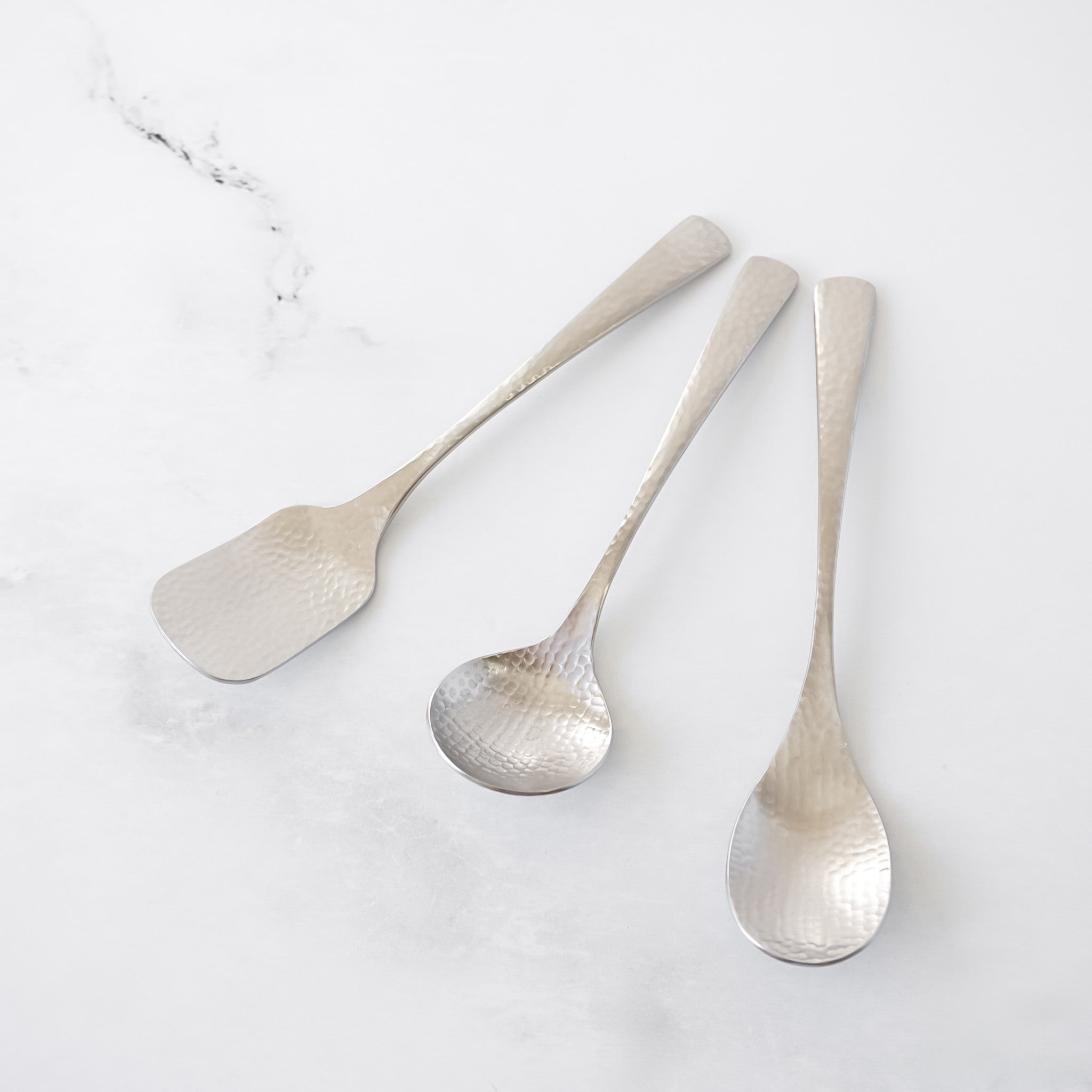 Three silver dessert spoons with a hammered texture. The set includes a ice cream spoon, a sugar spoon, and a teaspoon. The utensils are arranged on a white marble surface.
