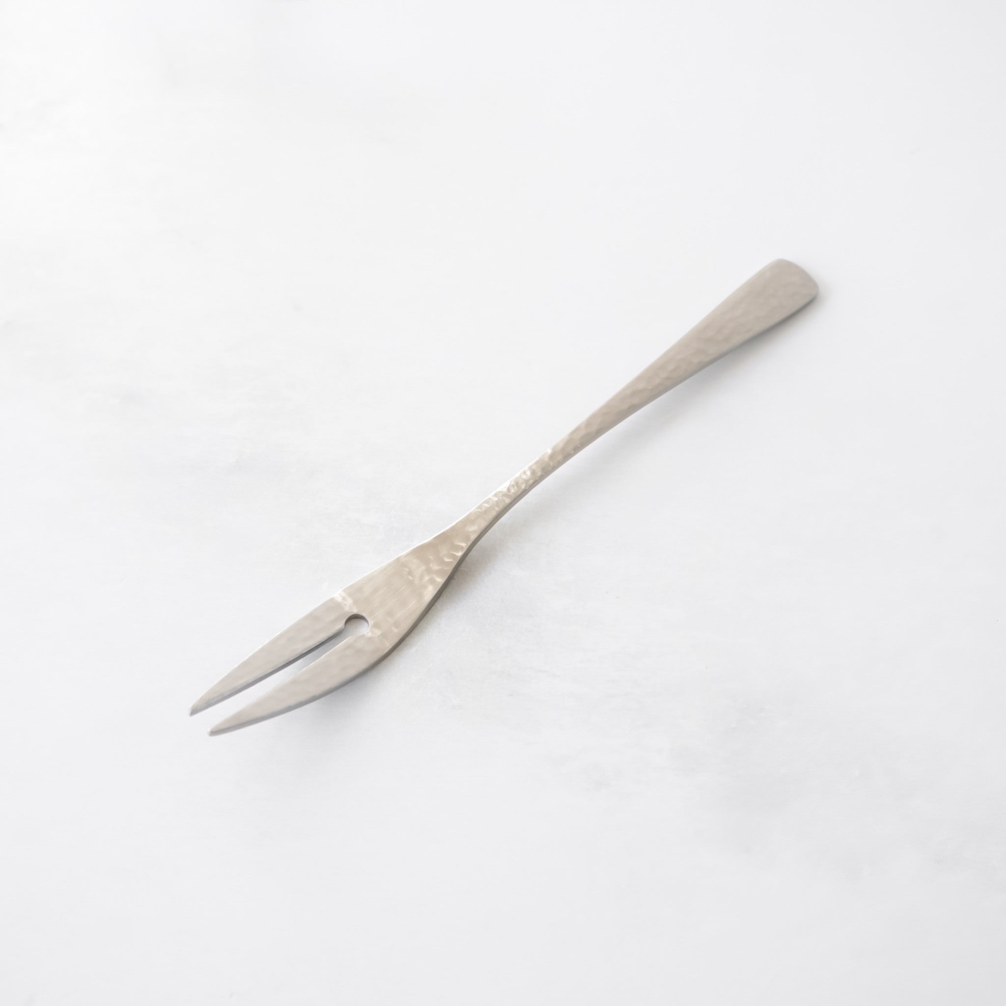 A single, silver-colored fork with two tines and a heart-shaped cutout. The handle is long and slender, with a textured surface. The fork is designed for serving appetizers or desserts.