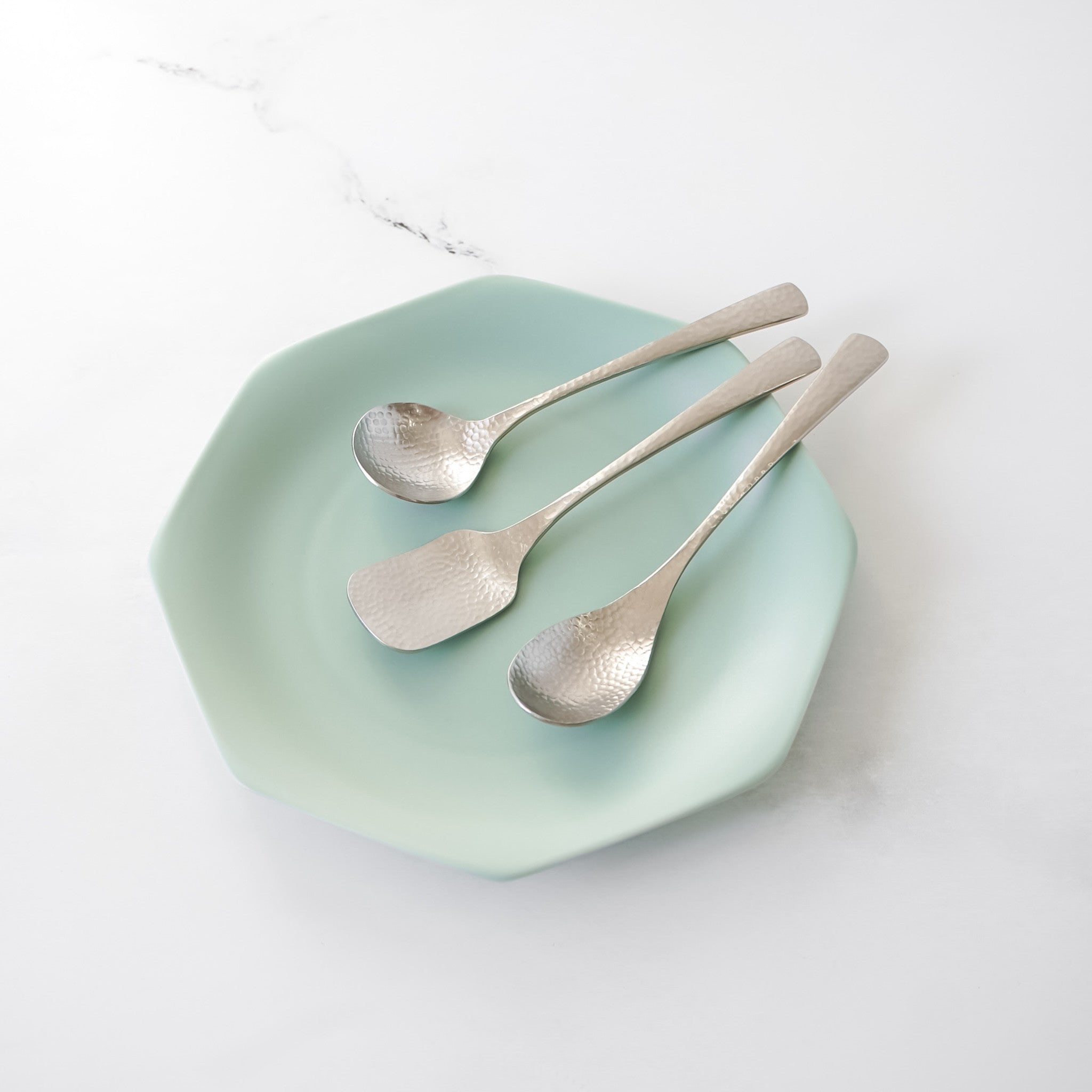 Three stainless steel dessert spoons with textured surfaces rest on a light green, octagonal plate. The spoons include a round spoon, a flat square spoon, and a teaspoon. The plate is set against a white marble background.