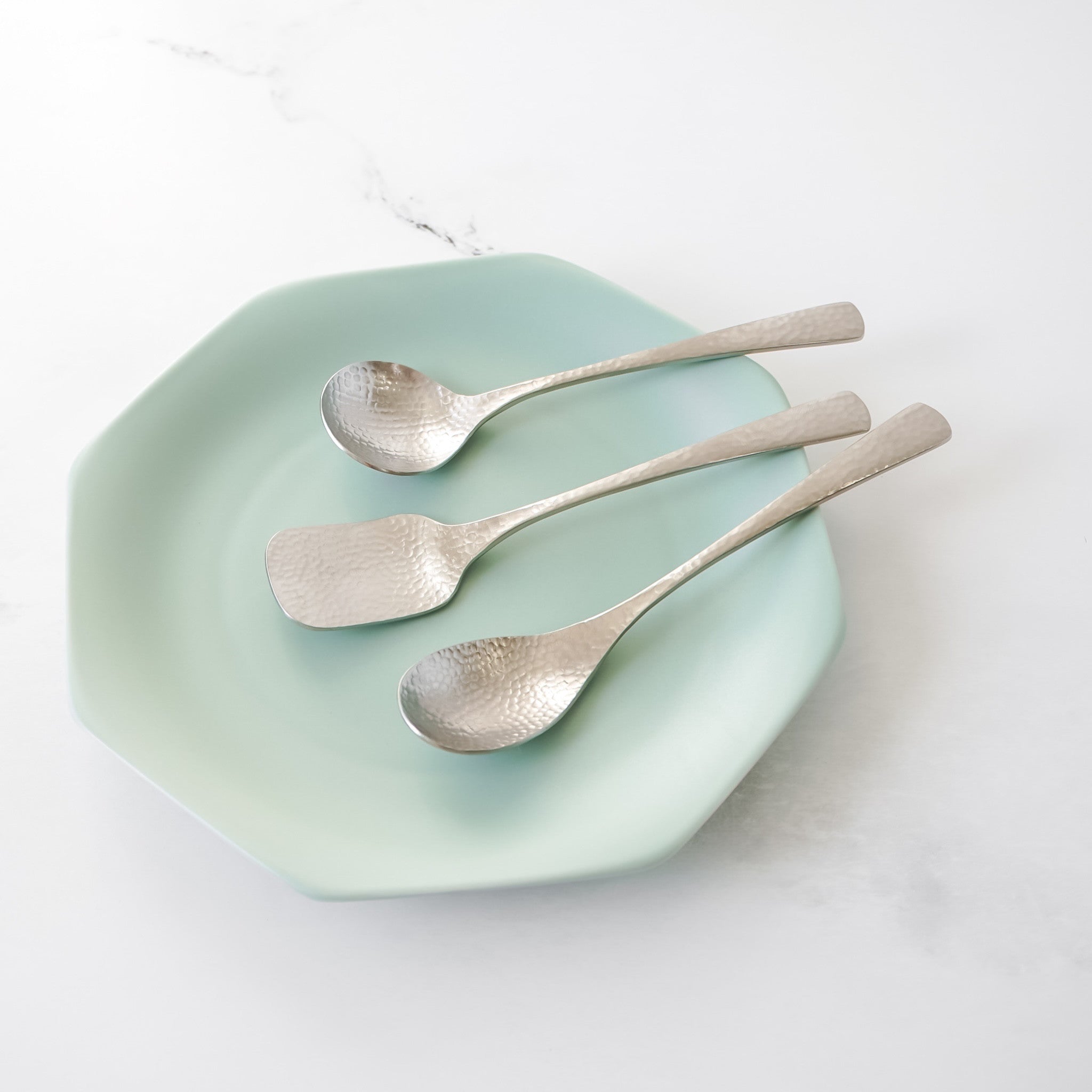 Three silver-colored dessert utensils with a hammered texture rest on a light green, octagonal plate. The utensils include three spoons. The plate is on a white surface.