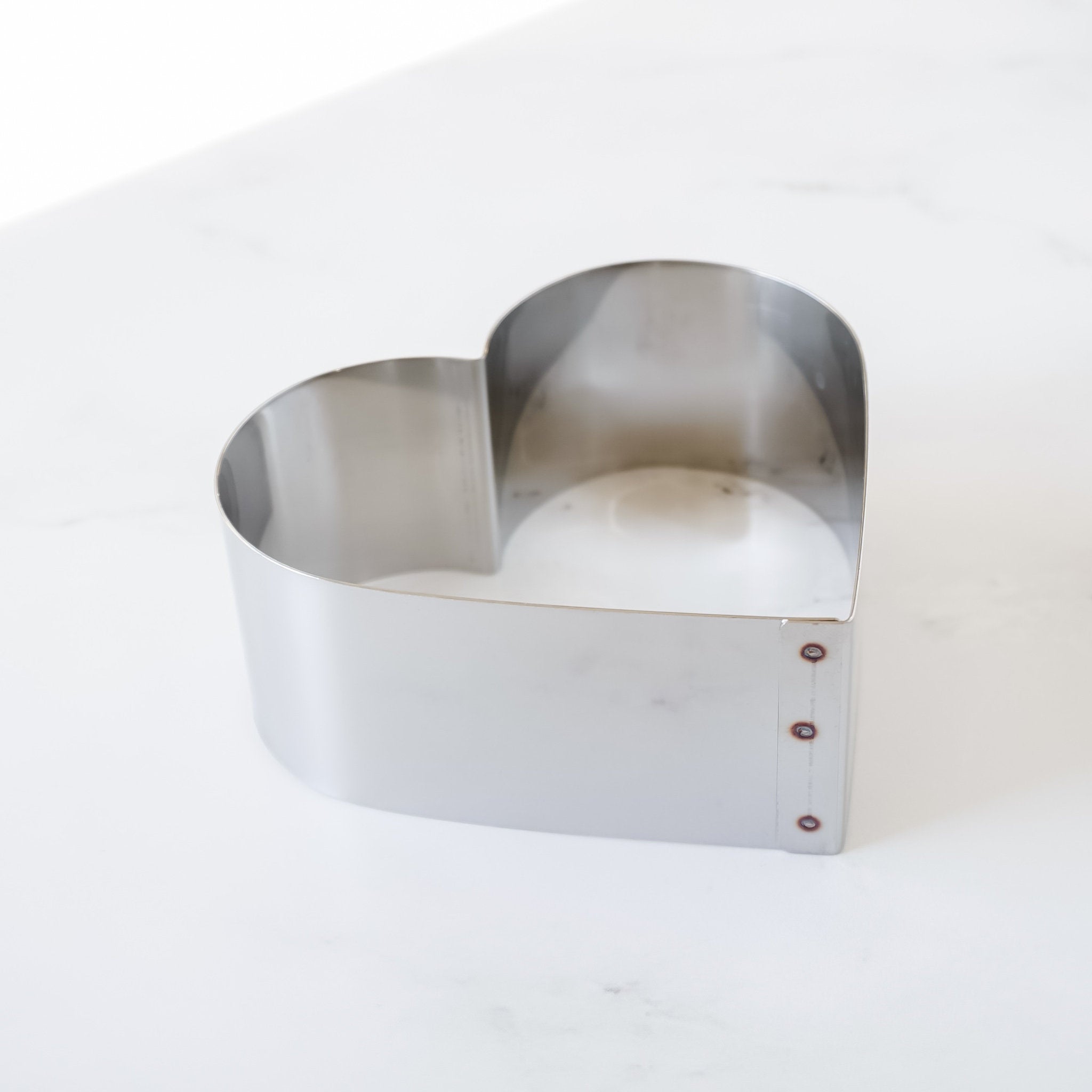 A silver heart-shaped cake pan without a bottom and a smooth, shiny finish. The pan has a seam on one side with three small, round rivets.