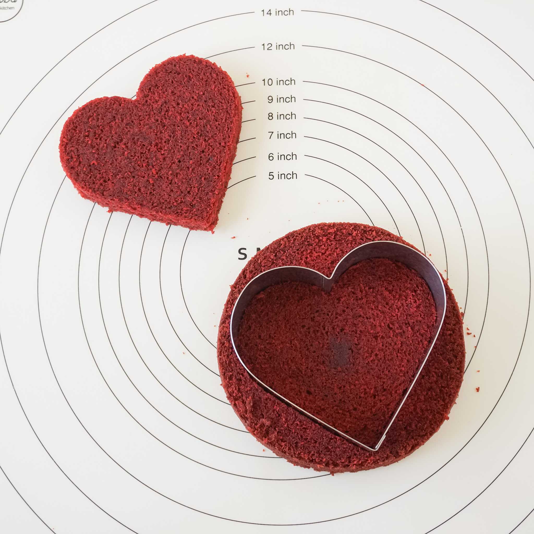 Two red velvet heart-shaped cakes on a white silicone baking mat with inch markings. The cake on the left is a whole heart and the cake on the right has a heart-shaped cake ring inside it.