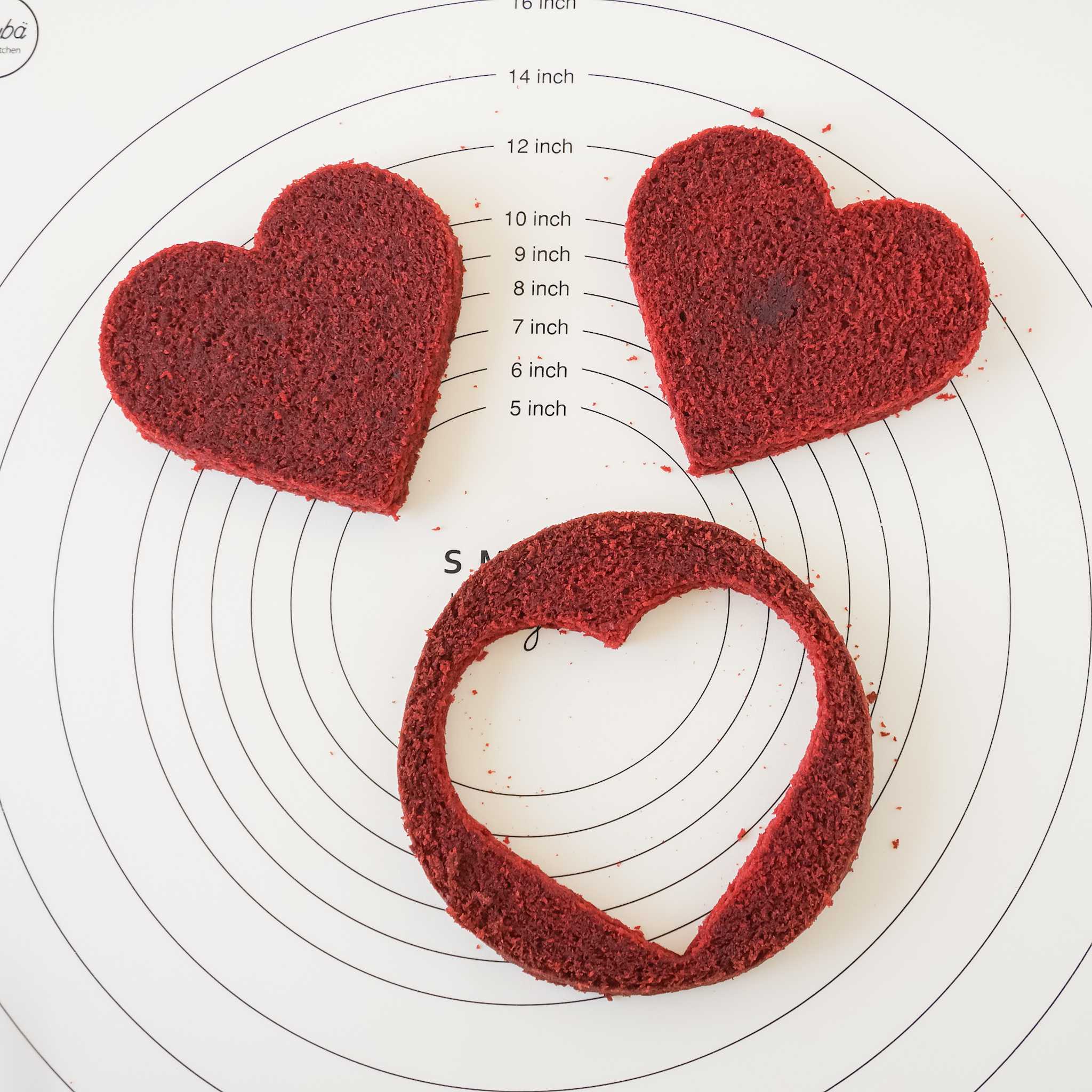 Three red velvet heart-shaped cakes on a white silicone baking mat with inch measurements. The cakes are arranged in a circle with the largest cake at the top and the smallest cake at the bottom. The cakes are all red velvet and have a smooth, even surface. 