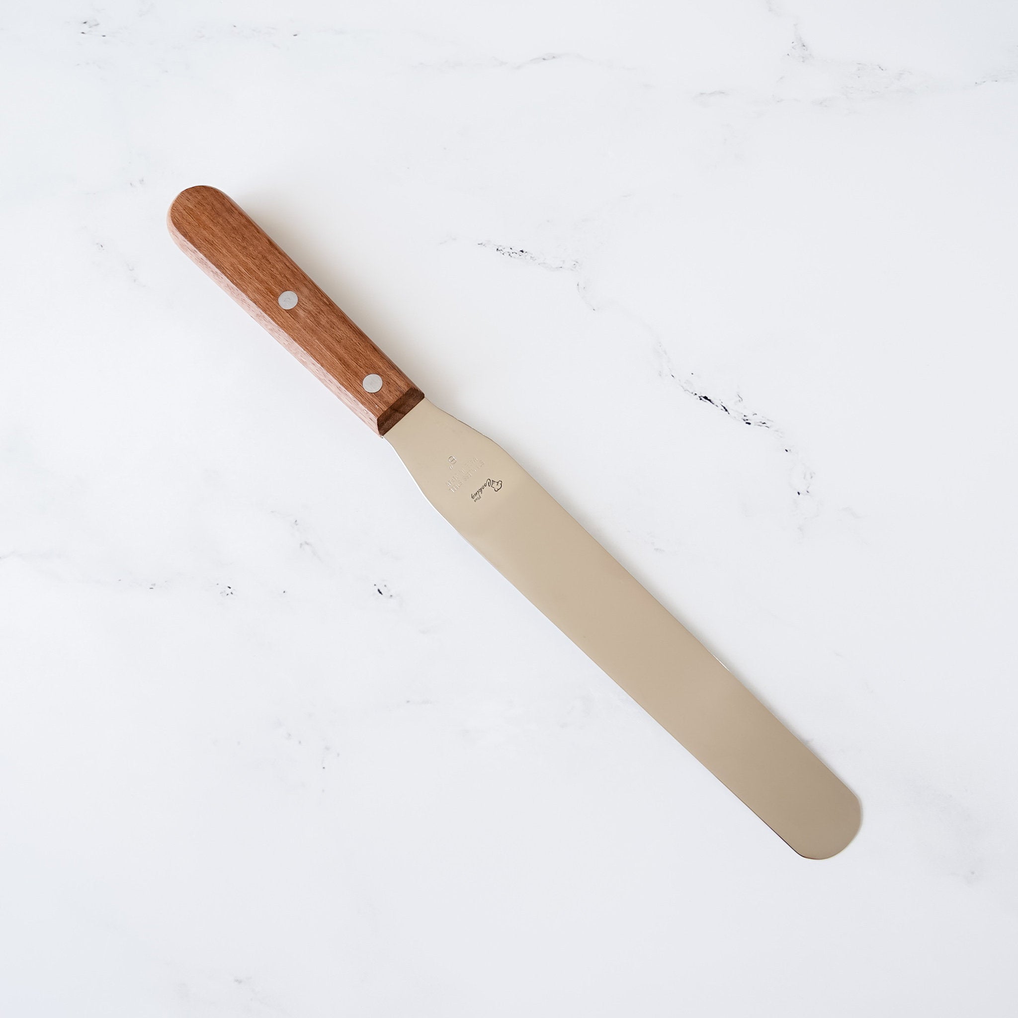 A stainless steel spatula with a wooden handle. The handle is brown and has two silver rivets. The spatula is on a white marble surface.