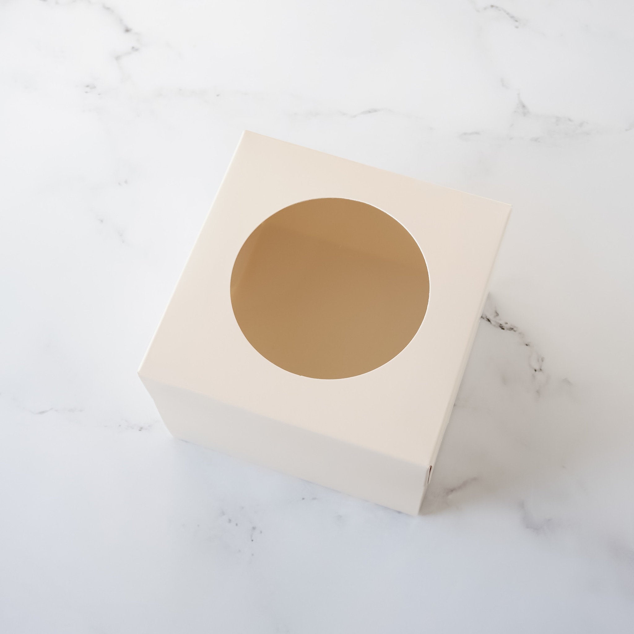  An ivory cardboard box with a round cutout in the center. The box is designed for packaging and displaying a single item, such as a cupcake or a small gift.