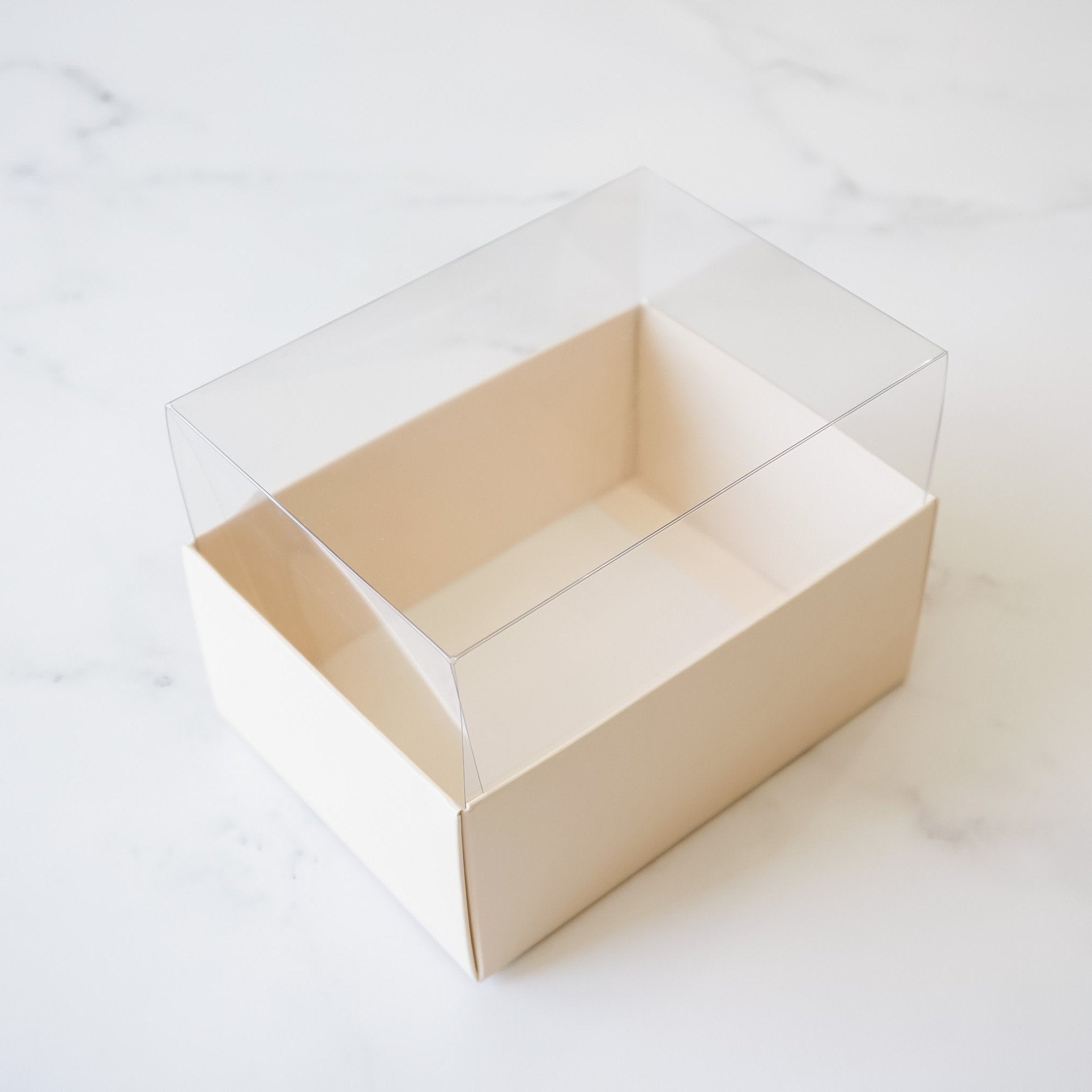 A rectangular cardboard box with a clear plastic lid. The box is an ivory color and is empty.