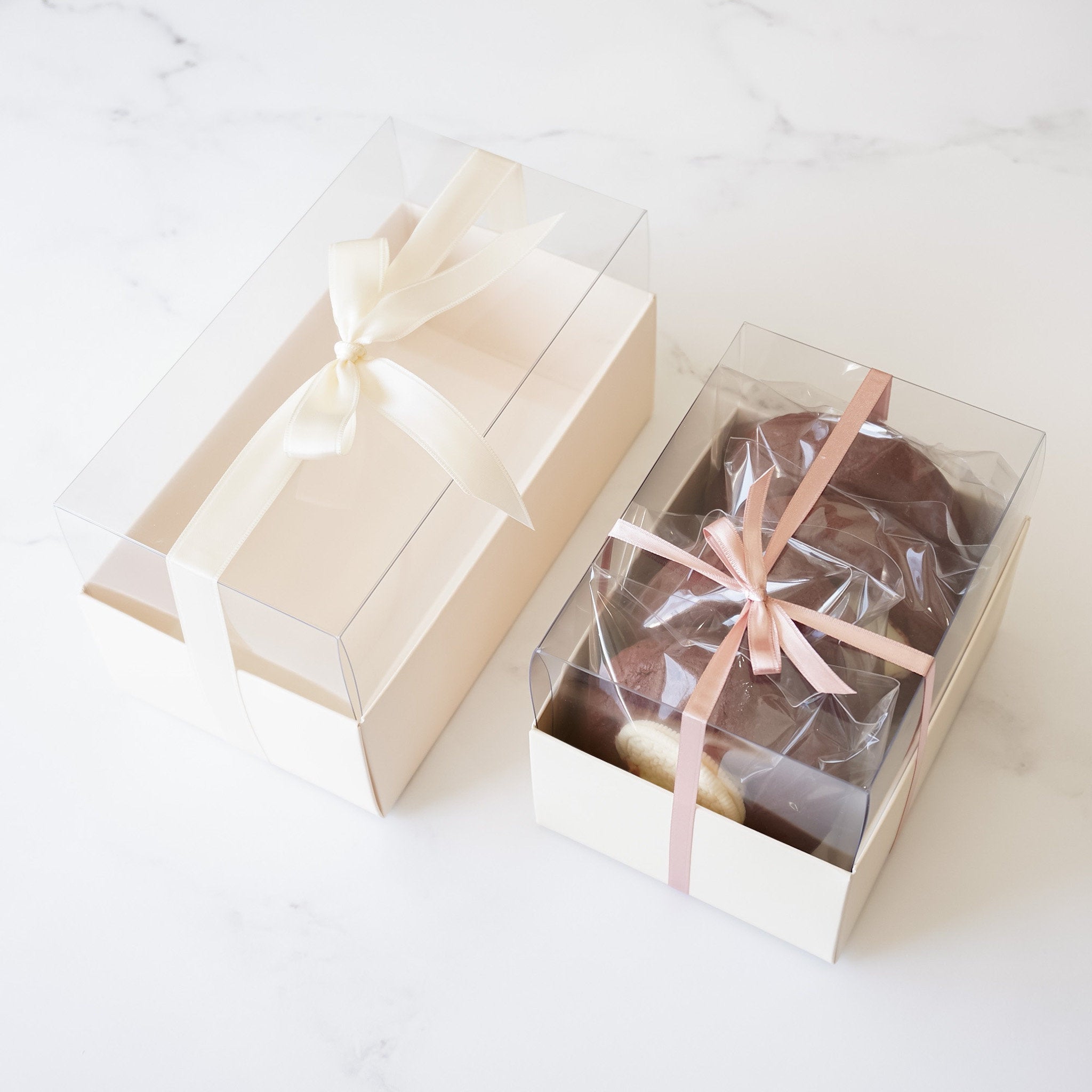 An ivory cookie box with a clear lid, open and tied with a satin bow, displaying a transparent wraping with cookies inside.