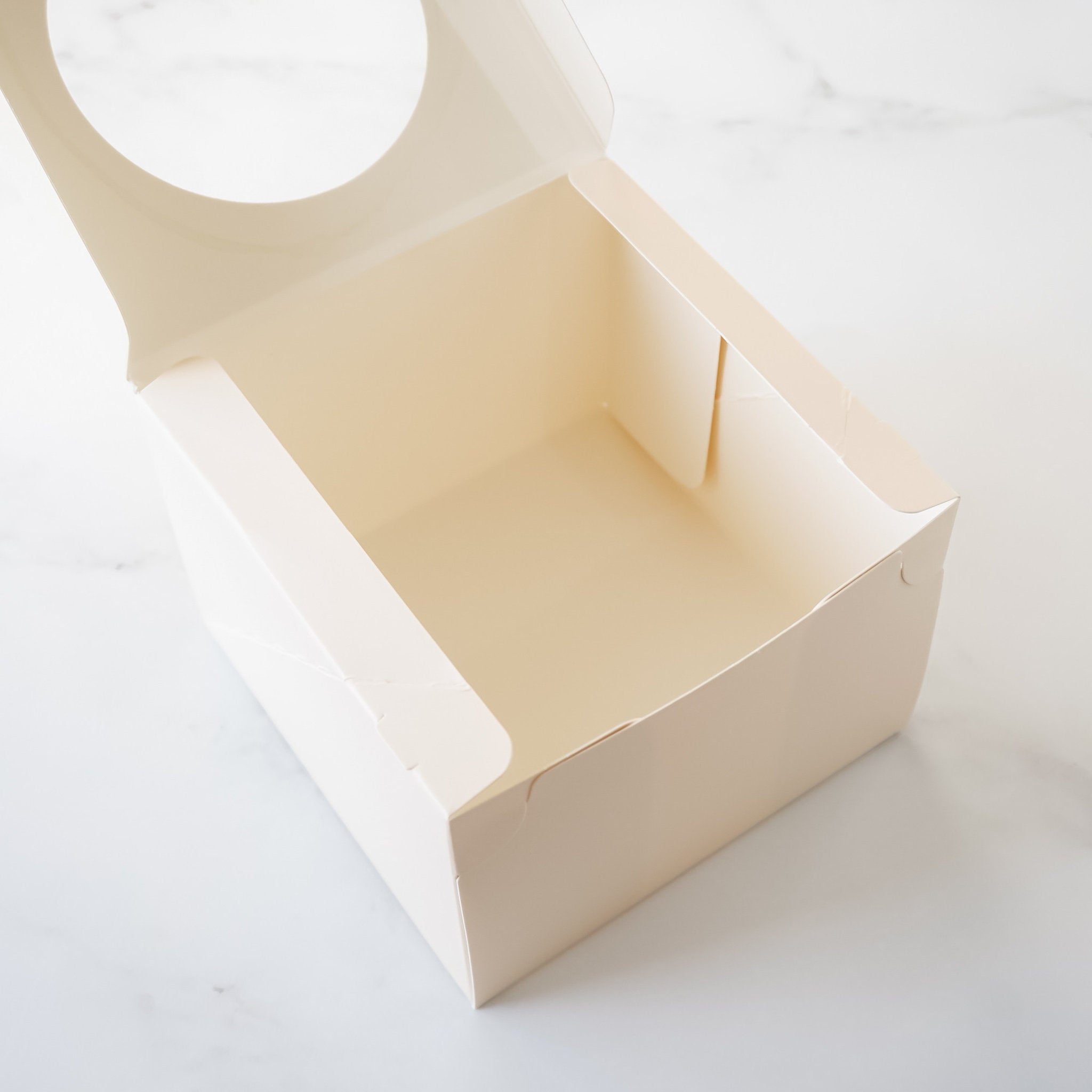 An empty, ivory cardboard box with a clear plastic window in the top. The box is open and the inside is visible. The box is sitting on a white marble surface.