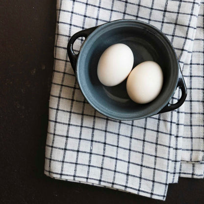 Two white eggs resting in a small dark gray ceramic pot with side handles, placed on a black-and-white checkered cloth over a dark surface.