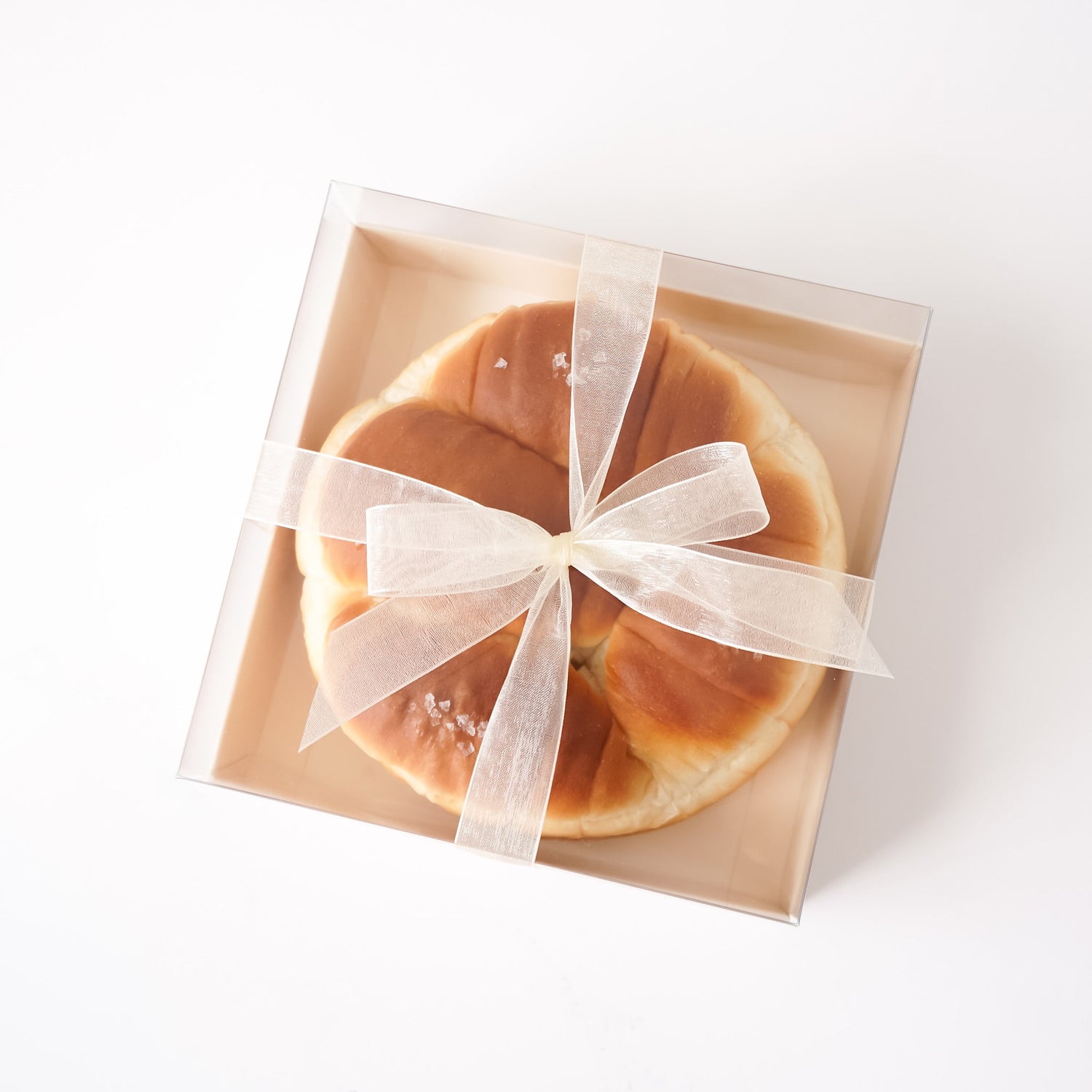 A round baked bun or pastry with a golden-brown crust and a few coarse salt crystals, presented inside a clear square gift box with a beige interior and tied with a sheer white ribbon.