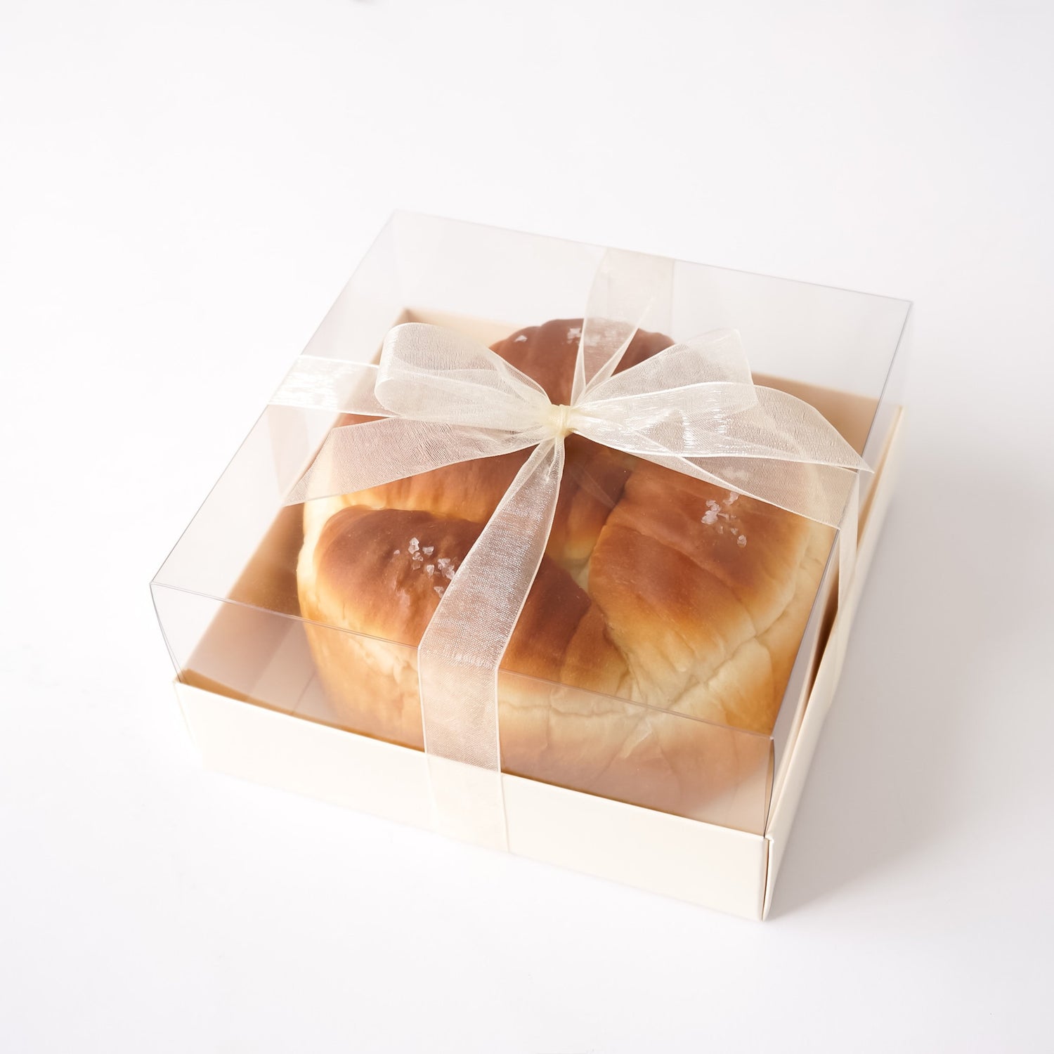 A round golden-brown bread roll, likely a brioche or sweet bread, presented inside a clear square plastic bakery box with a white organza ribbon tied in a bow, on a clean white background.