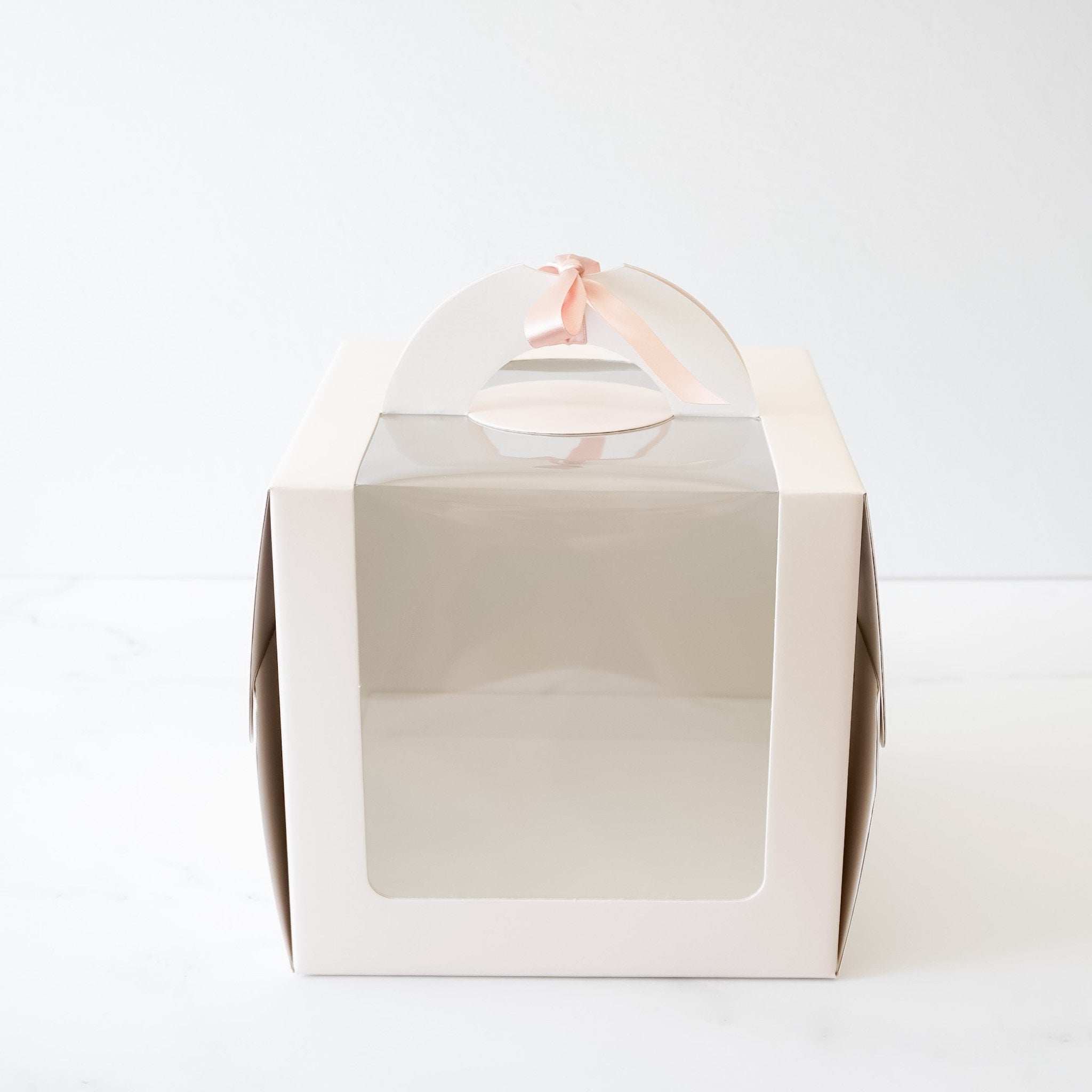 A white cake with white frosting in a clear plastic box with a white cardboard lid and a pink ribbon handle.