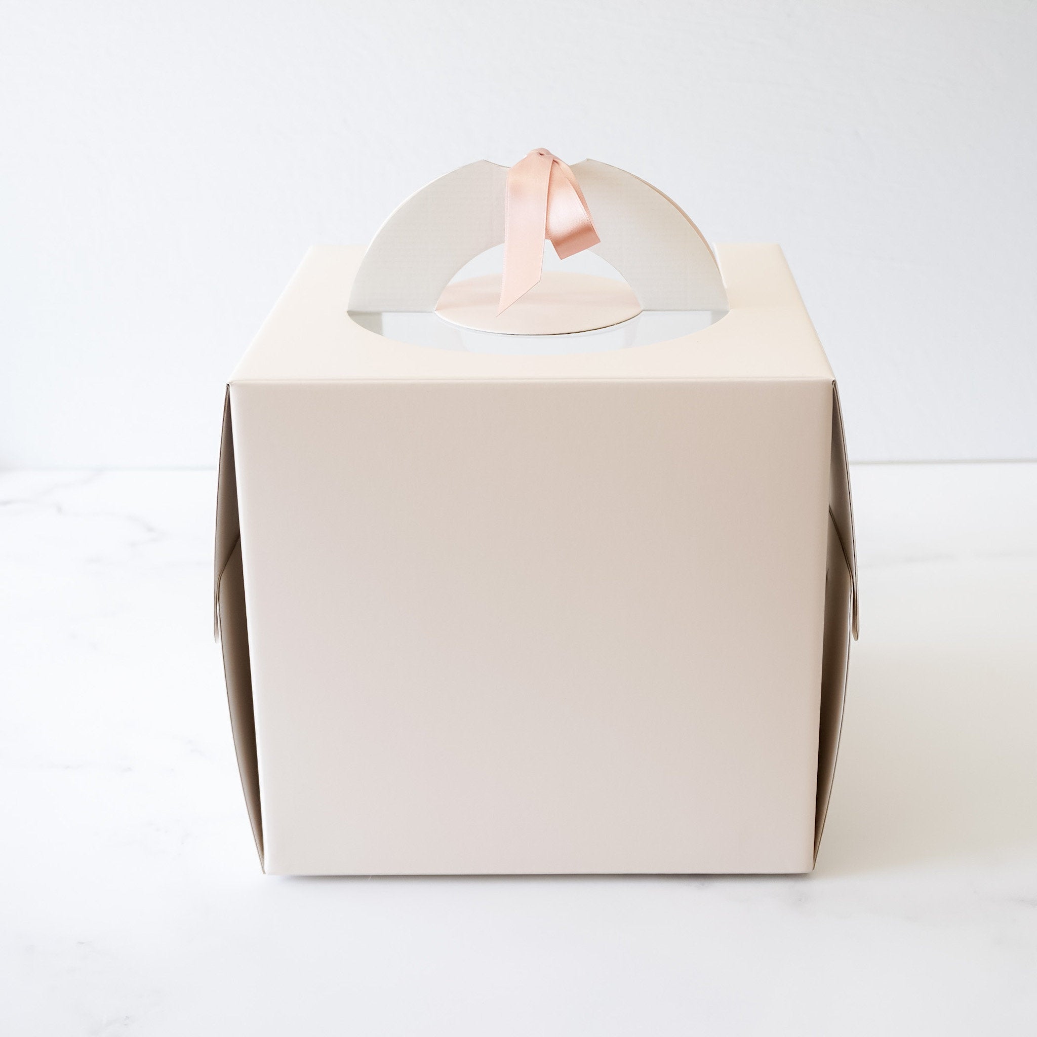A white cardboard cake box with a clear plastic window on the top. The box has a handle made of a white cardboard arch with a pink satin ribbon attached to it. The box is sitting on a white marble surface.