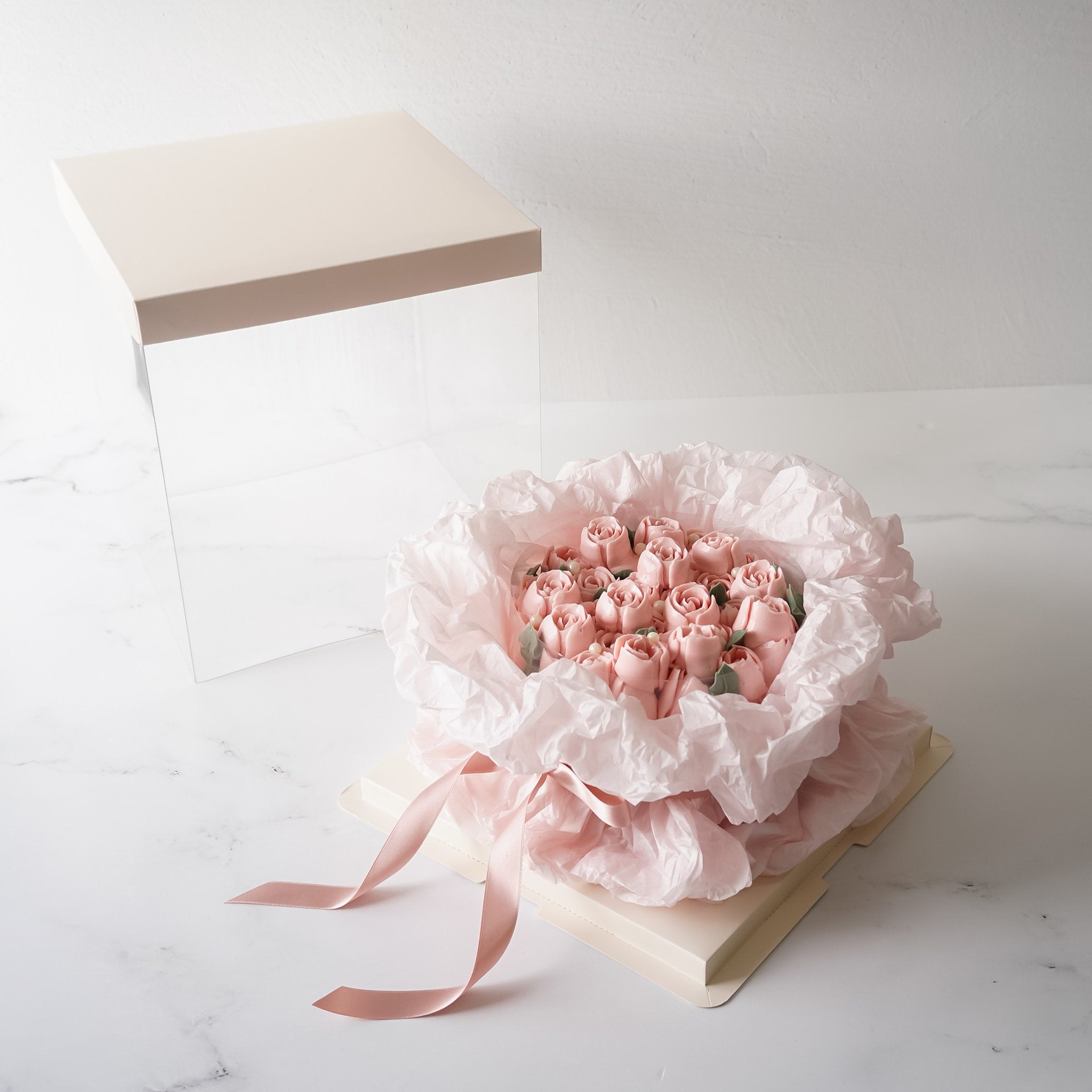 A pink rose bouquet cake wrapped in white tissue paper and tied with a pink ribbon. The bouquet is sitting on a white cardboard box with a clear acrylic lid.