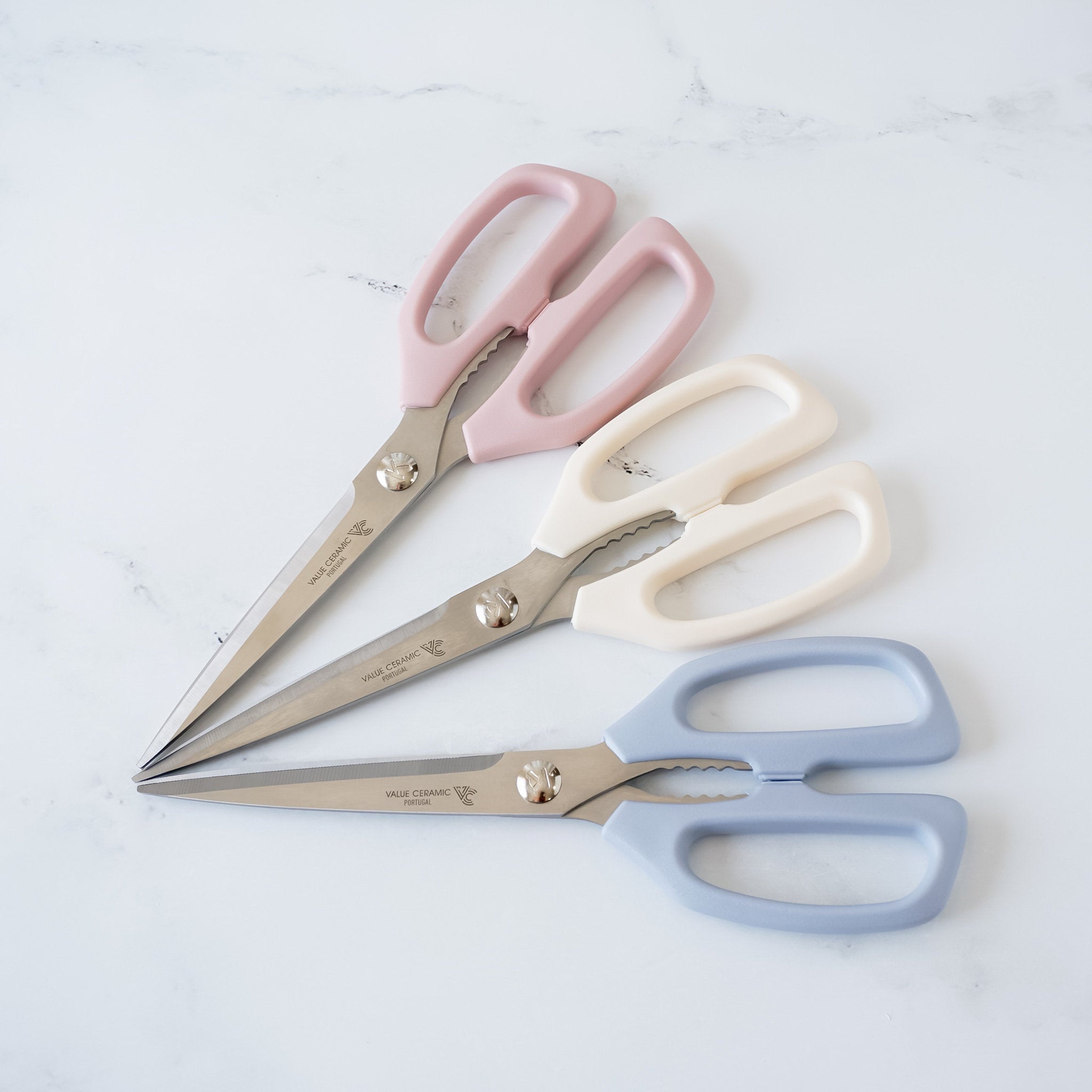 Three pairs of kitchen shears with ceramic blades. The shears are in pink, white, and blue. The blades are stainless steel and have a serrated edge. The handles are made of plastic and have a comfortable grip. 