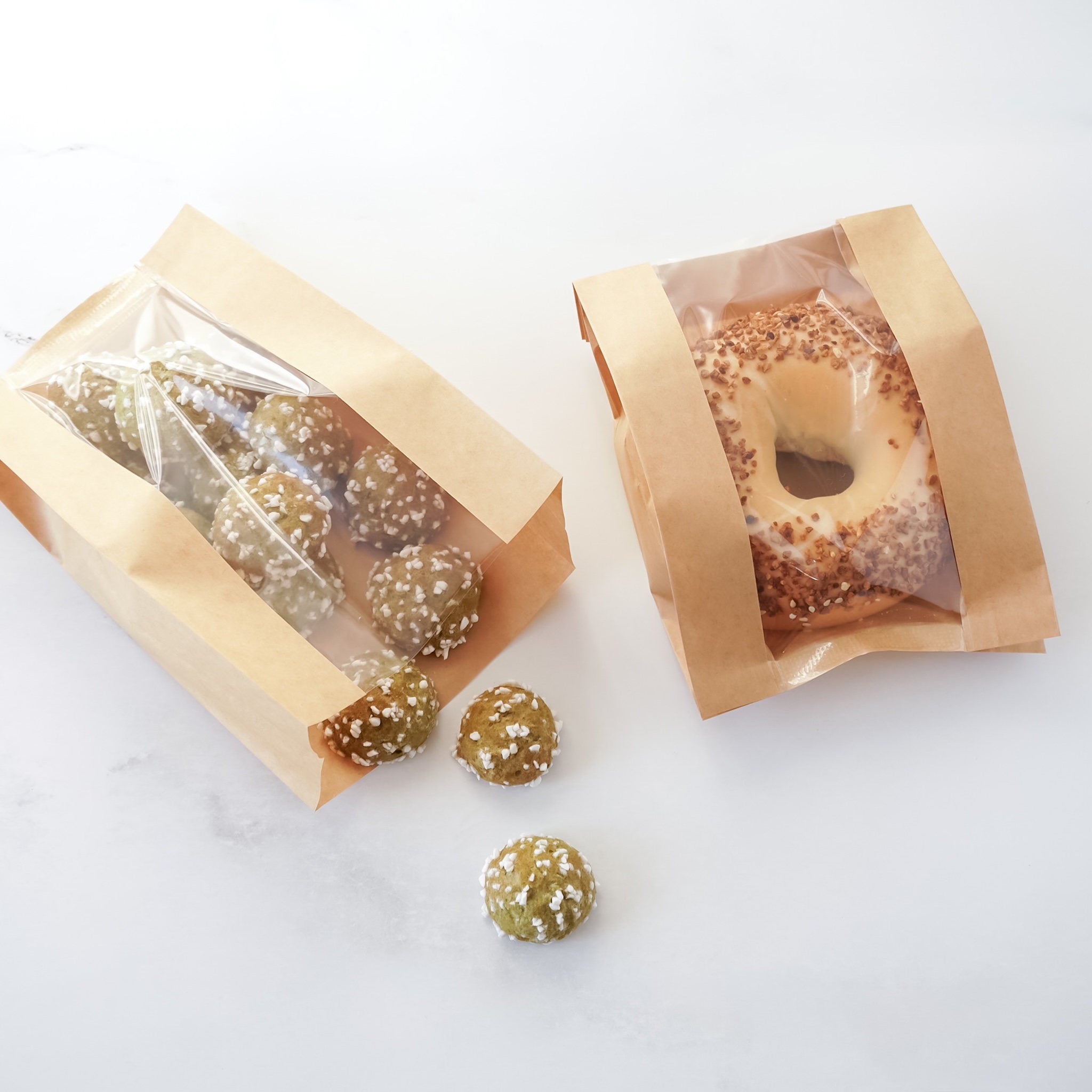 Two brown paper bags with clear window displays containing round, textured snacks on a white background.