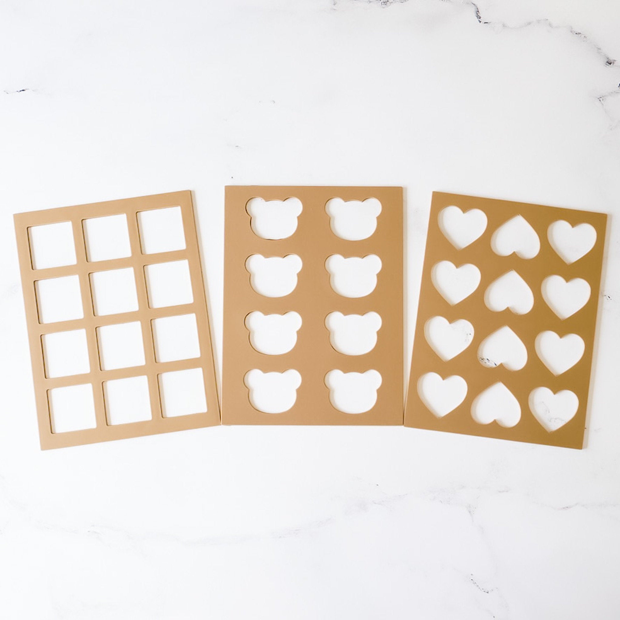 Three silicone cookie stencils with shapes for squares, hearts, and a bear, laid out on a white surface.