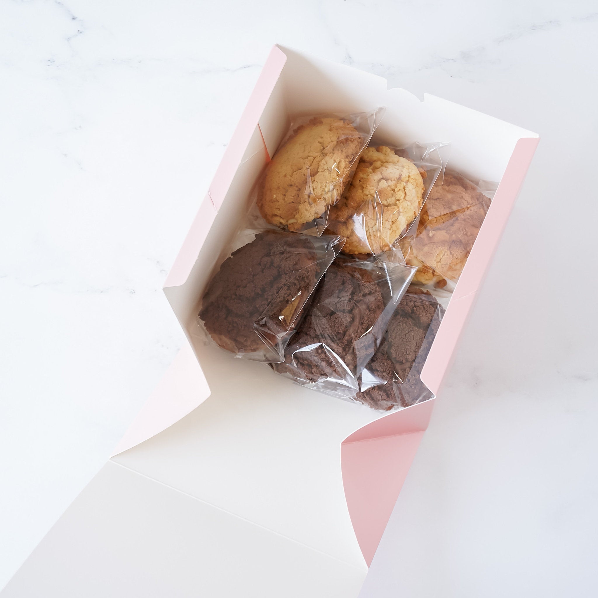 A pink box with a white interior containing six cookies. Three cookies are light brown and three cookies are dark brown. The cookies are wrapped in clear plastic and are arranged in a box.