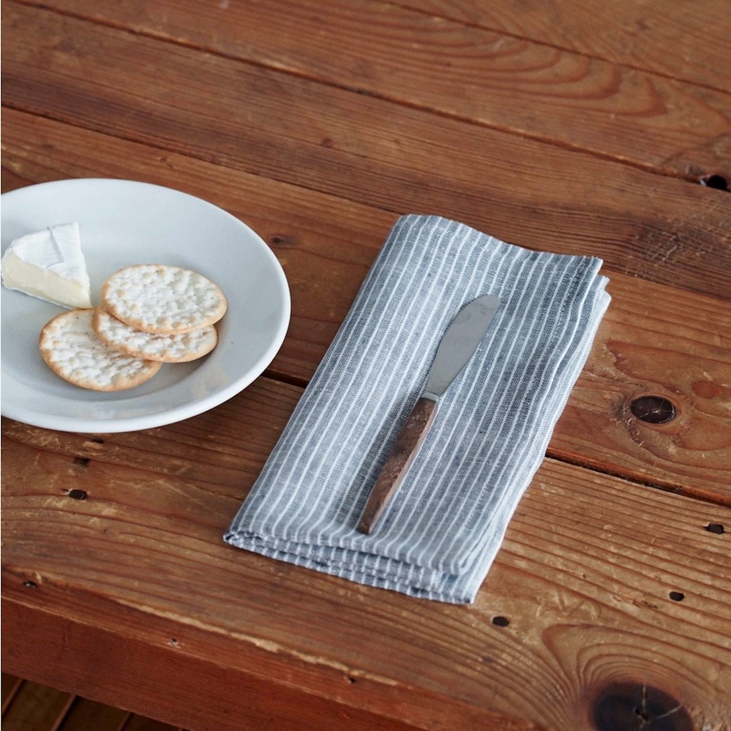 Napkin with a knife on a wooden table next to a plate of cheese and crackers