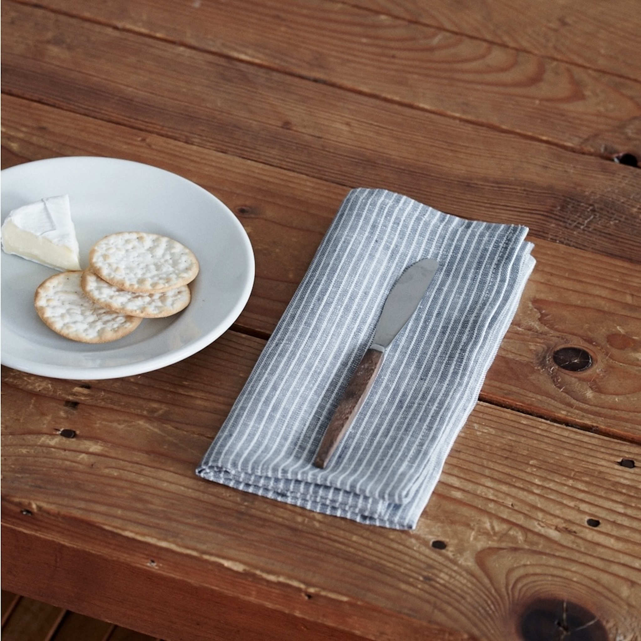 Napkin with a knife on a wooden table next to a plate of cheese and crackers