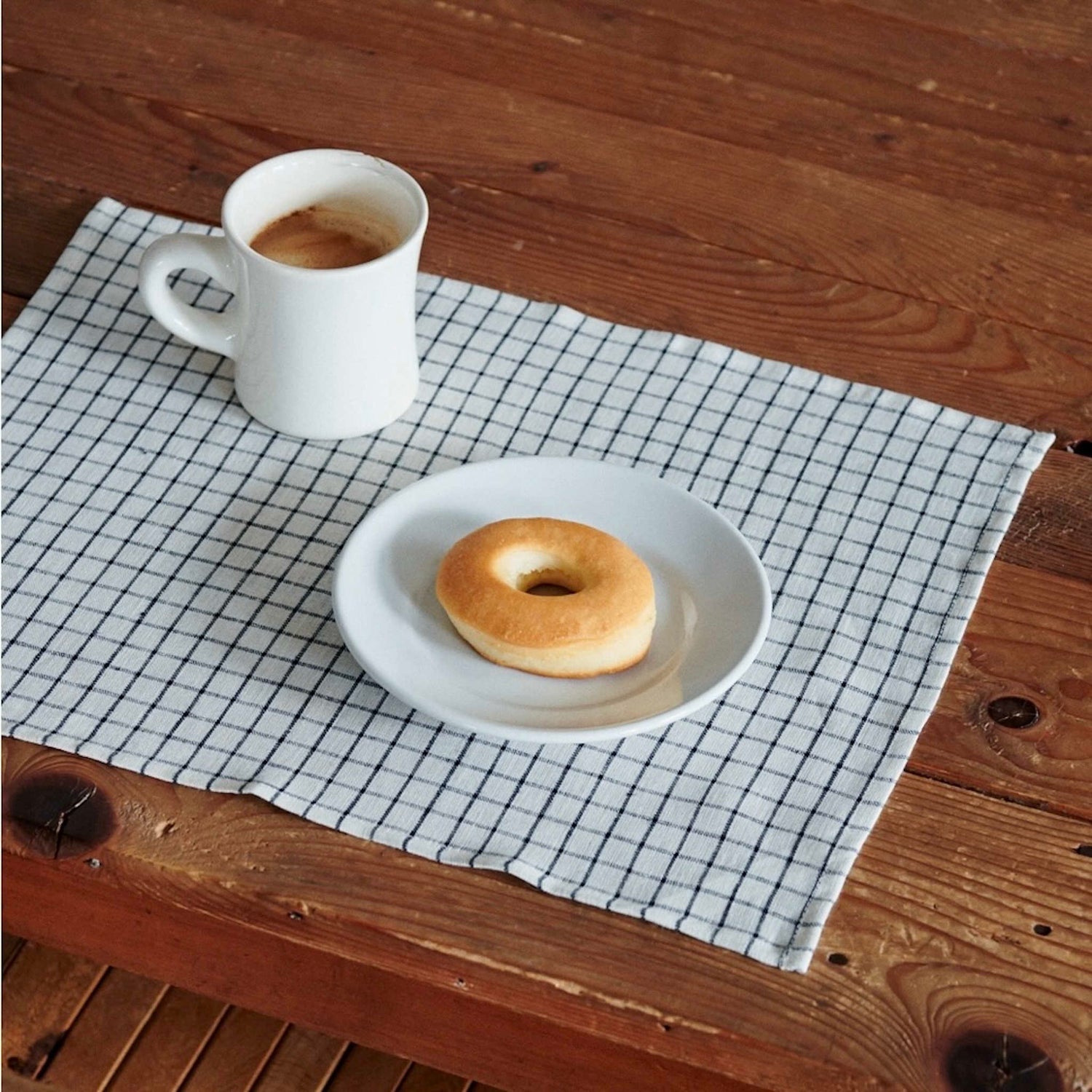 White mug with coffee and a donut on a white plate on a checkered table placemat over a wooden table.
