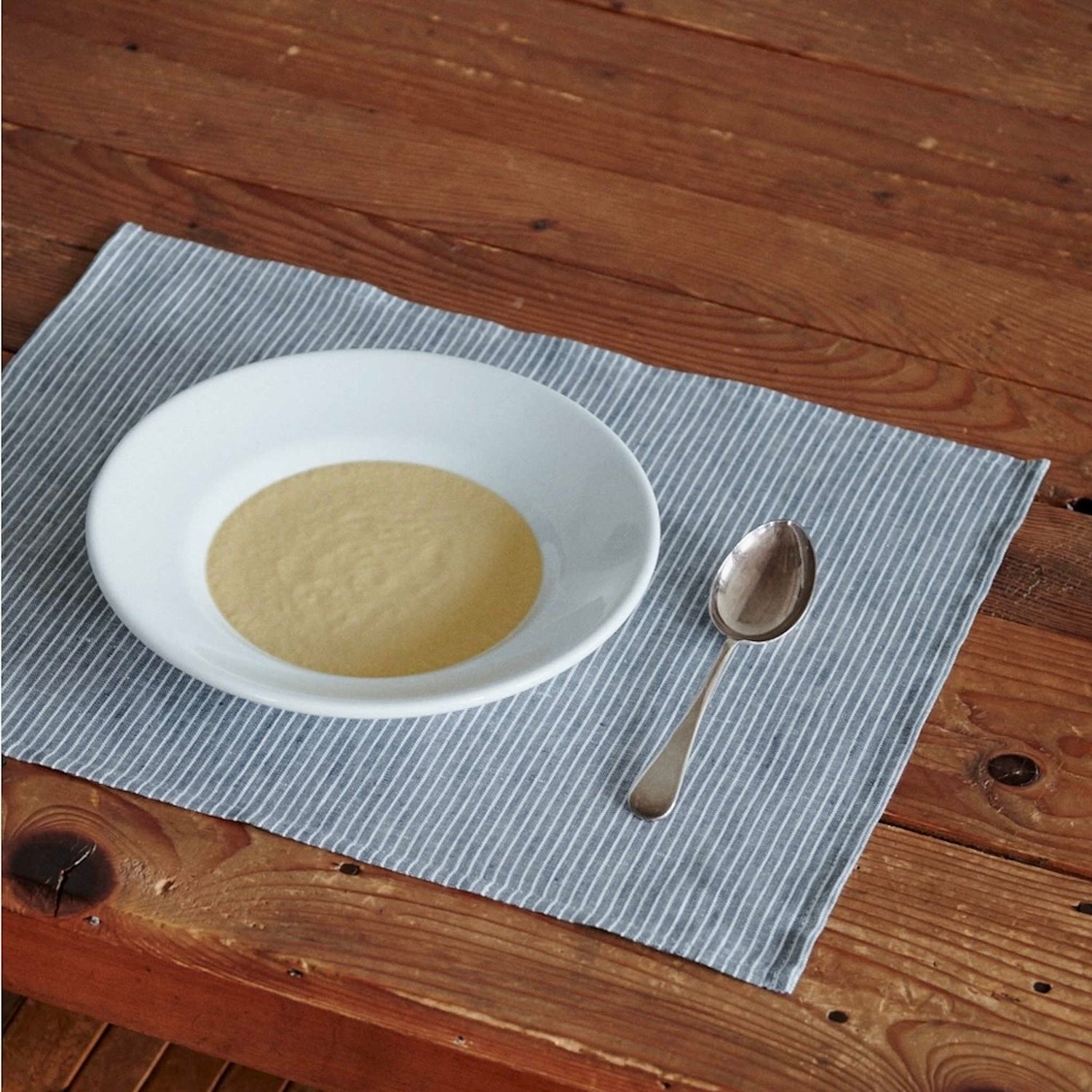White plate with soup and a spoon on a gray striped placemat on a wooden table