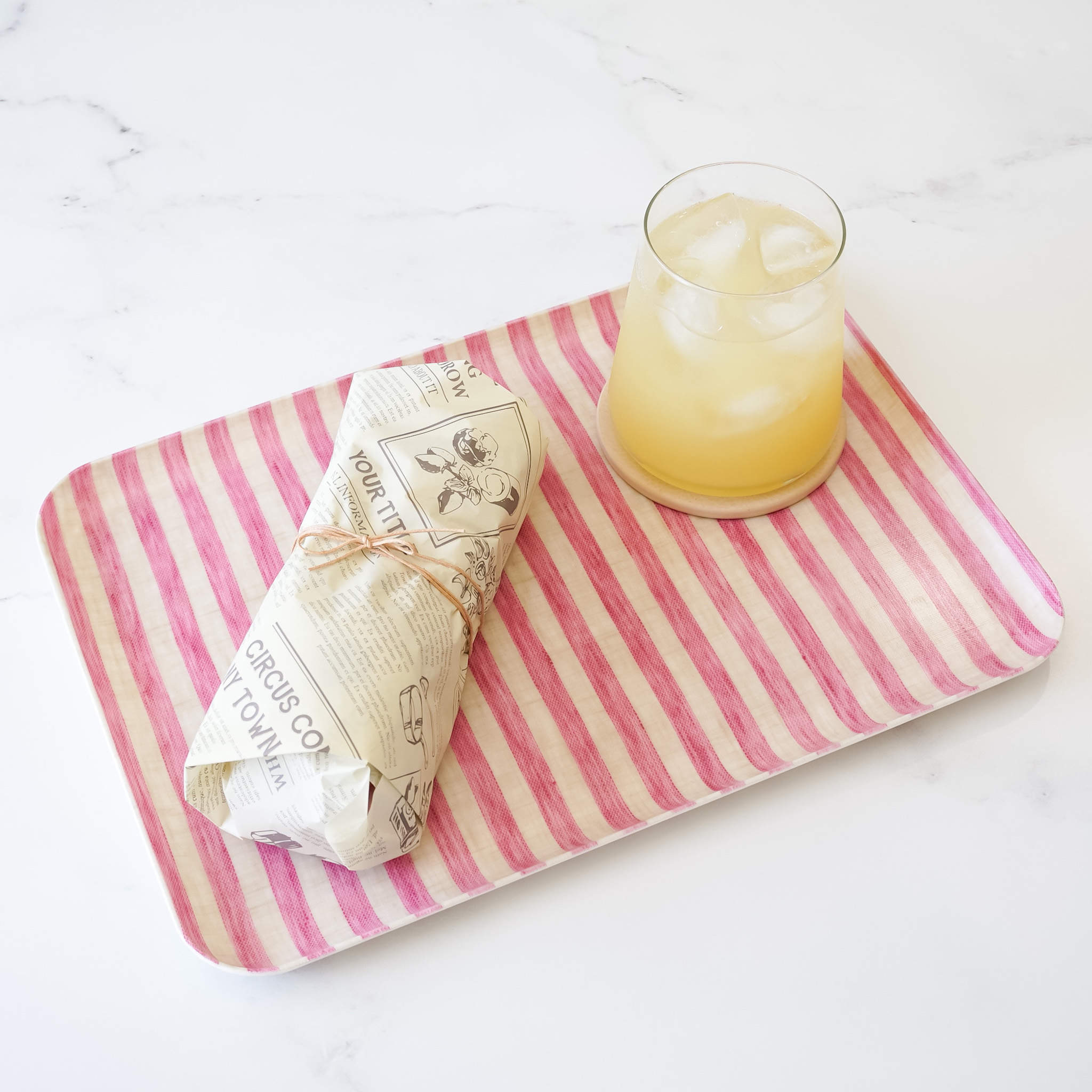 A pink striped linen coated tray with a glass of iced drink and a wrapped sandwich on a marble surface.