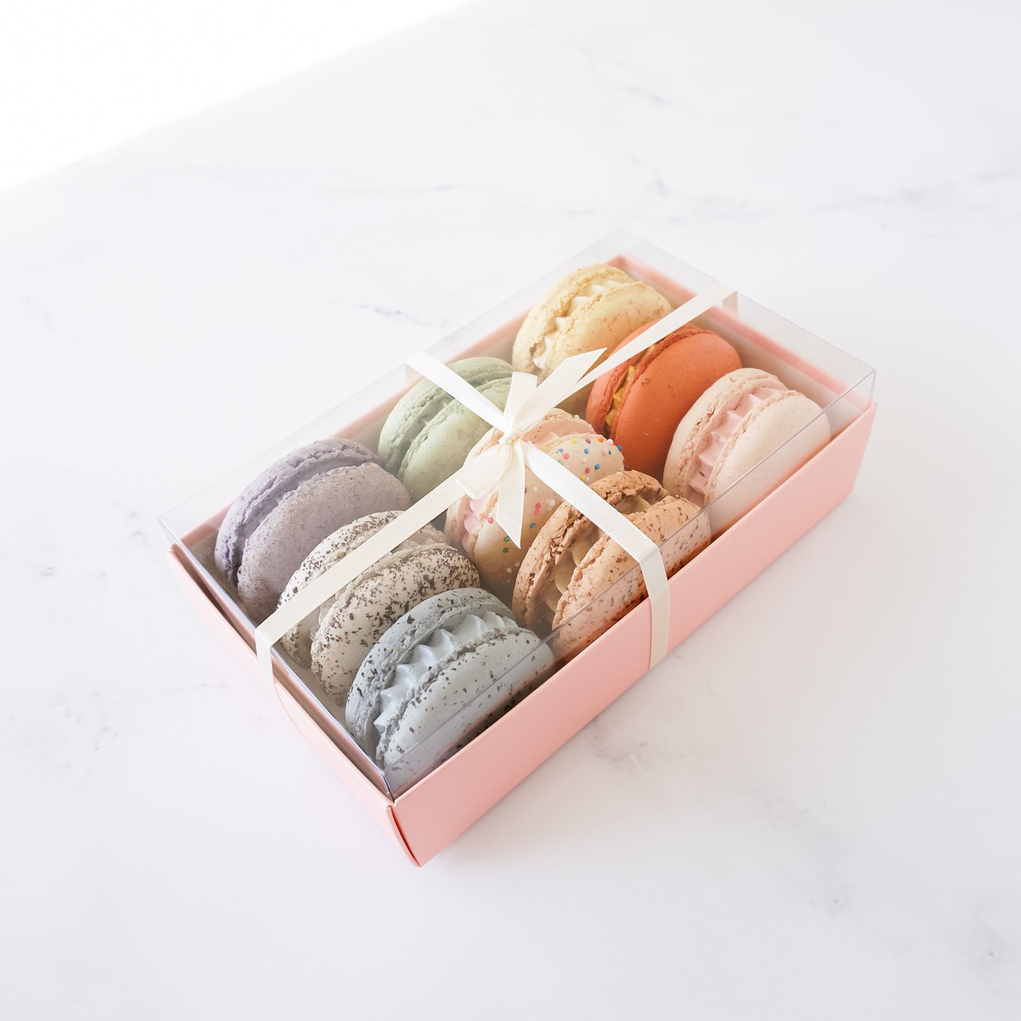 A pink box filled with colorful macarons. The macarons are arranged in a circle with the pink box in the center. The macarons are a variety of colors, including pink, green, blue, and yellow. The macarons are a popular French dessert.