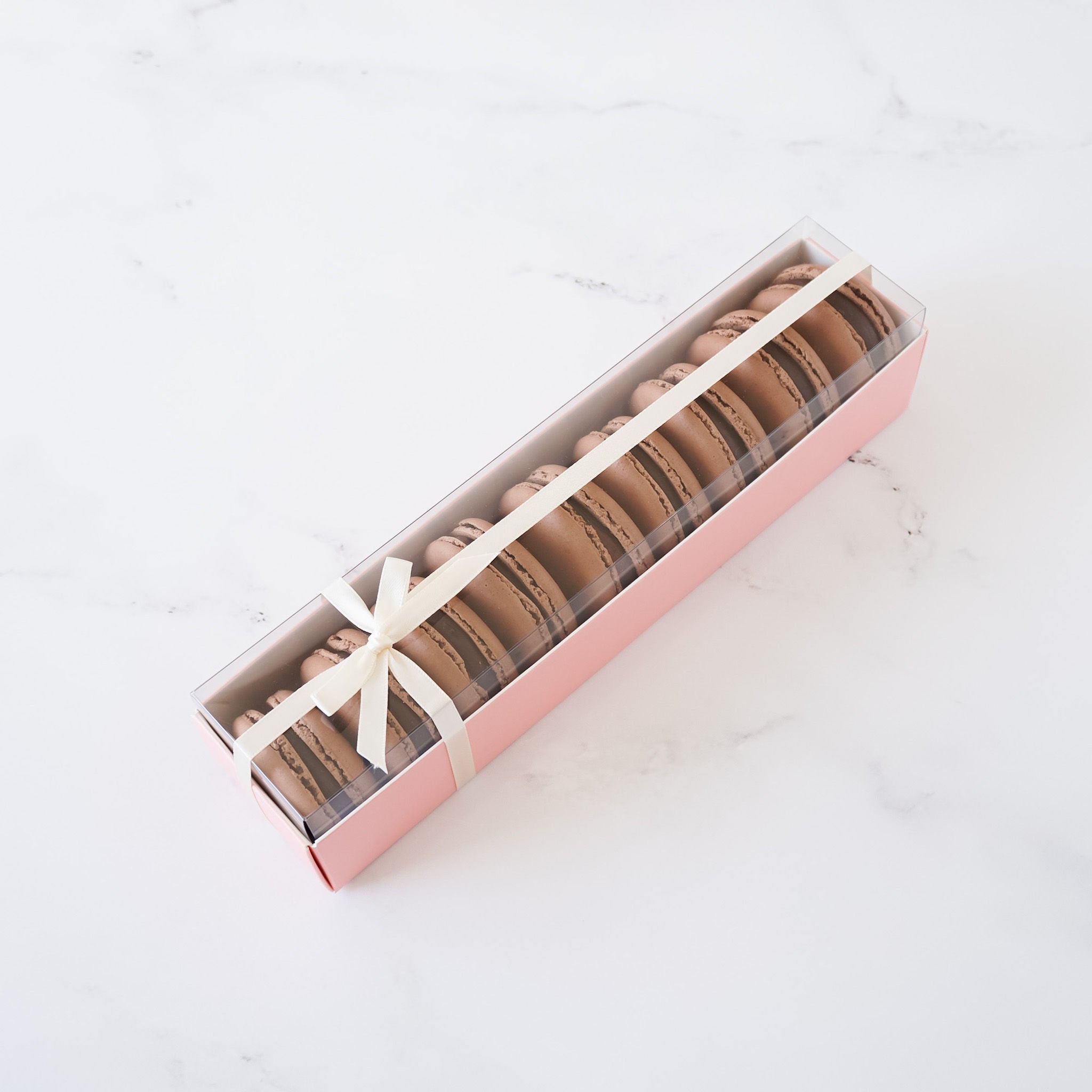 A pink box with a clear lid filled with a row of chocolate macarons tied with a white ribbon.
