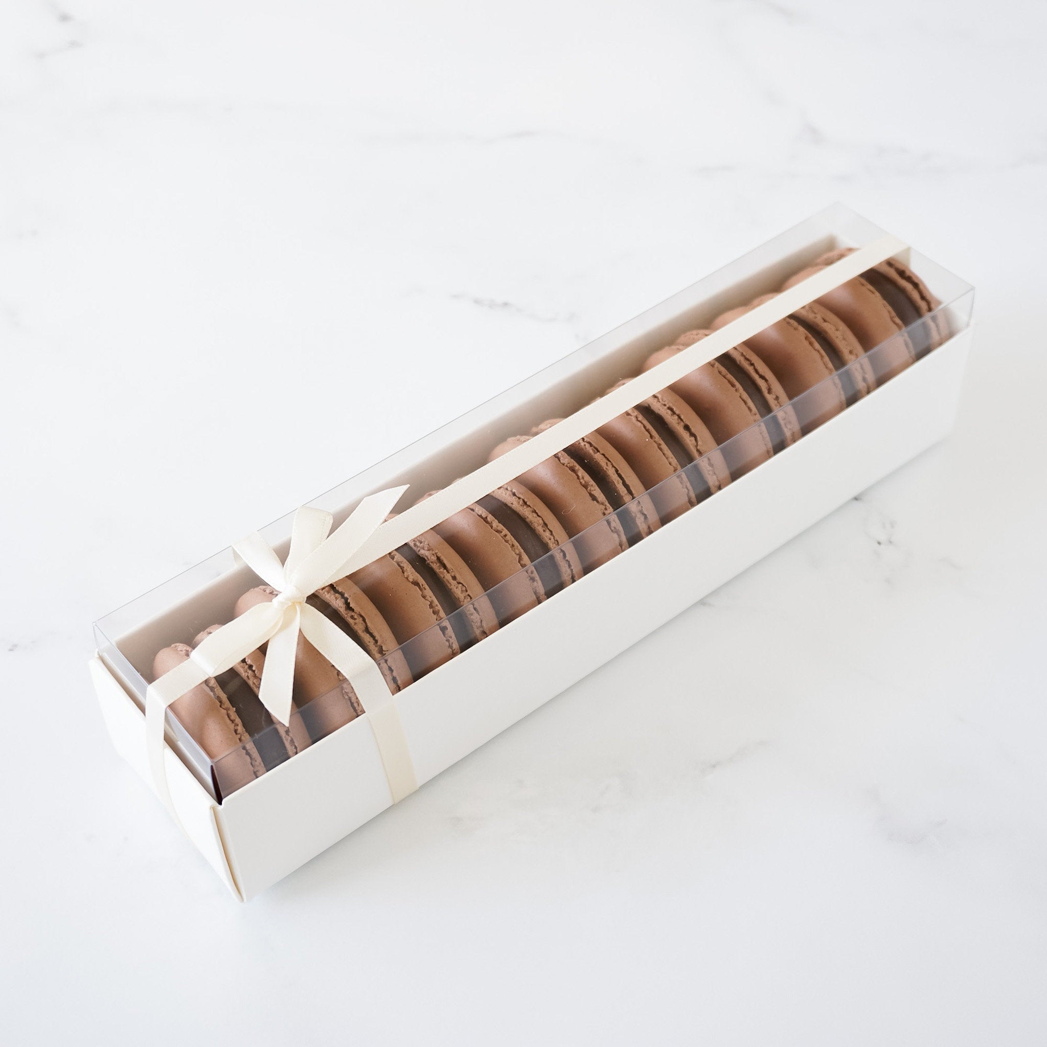 A white box with a white ribbon tied around it. The box is filled with chocolate macarons. The macarons are arranged in a row, with the top of each macaron visible. The box is sitting on a white surface.