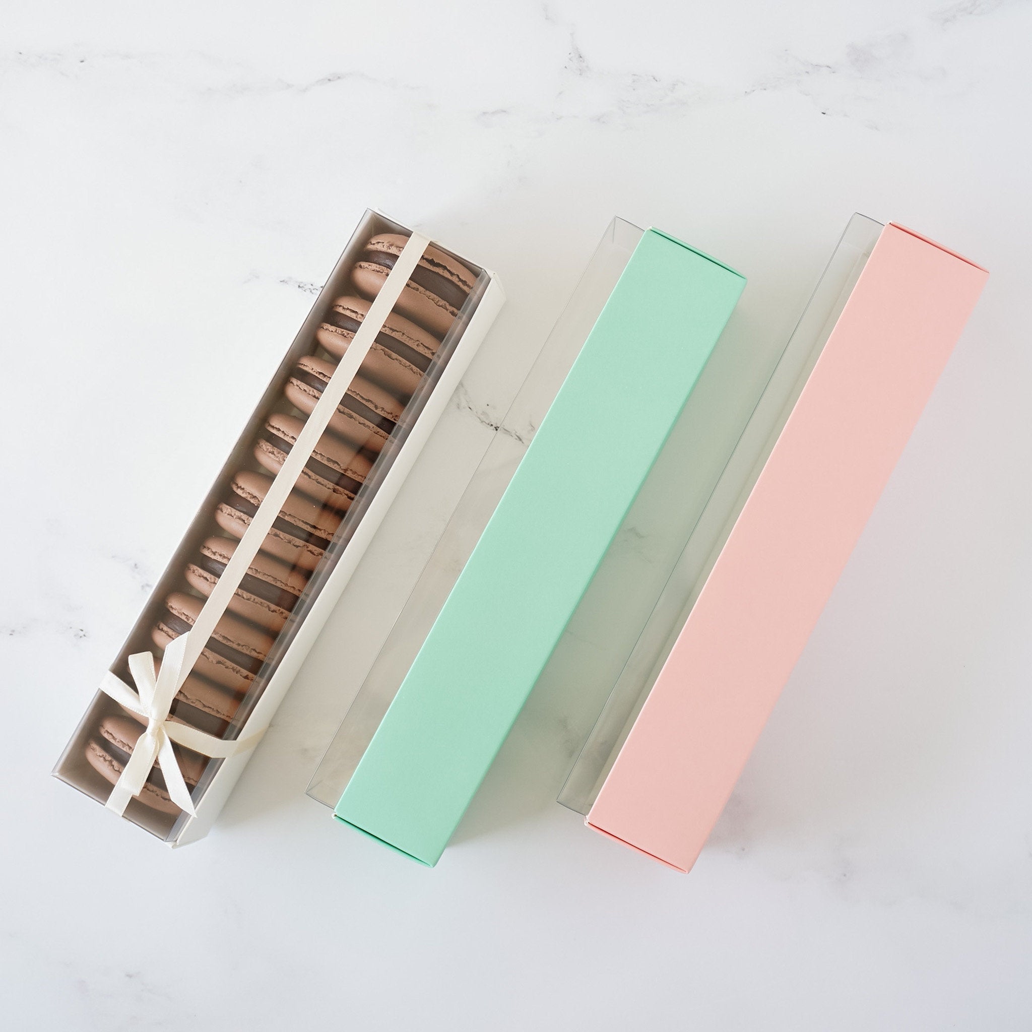 Three rectangular boxes with clear lids. The box on the left contains a row of chocolate macarons. The middle box is light green and the box on the right is light pink.