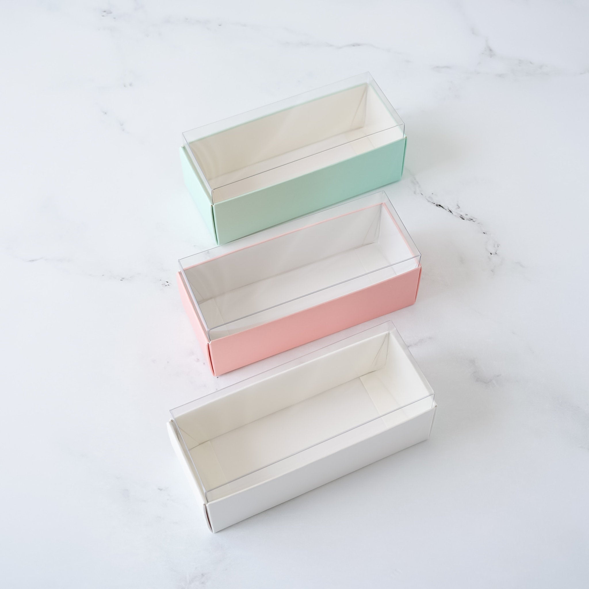 Three rectangular boxes with clear plastic lids. The boxes are stacked on top of each other. The top box is light green, the middle box is pink, and the bottom box is white.