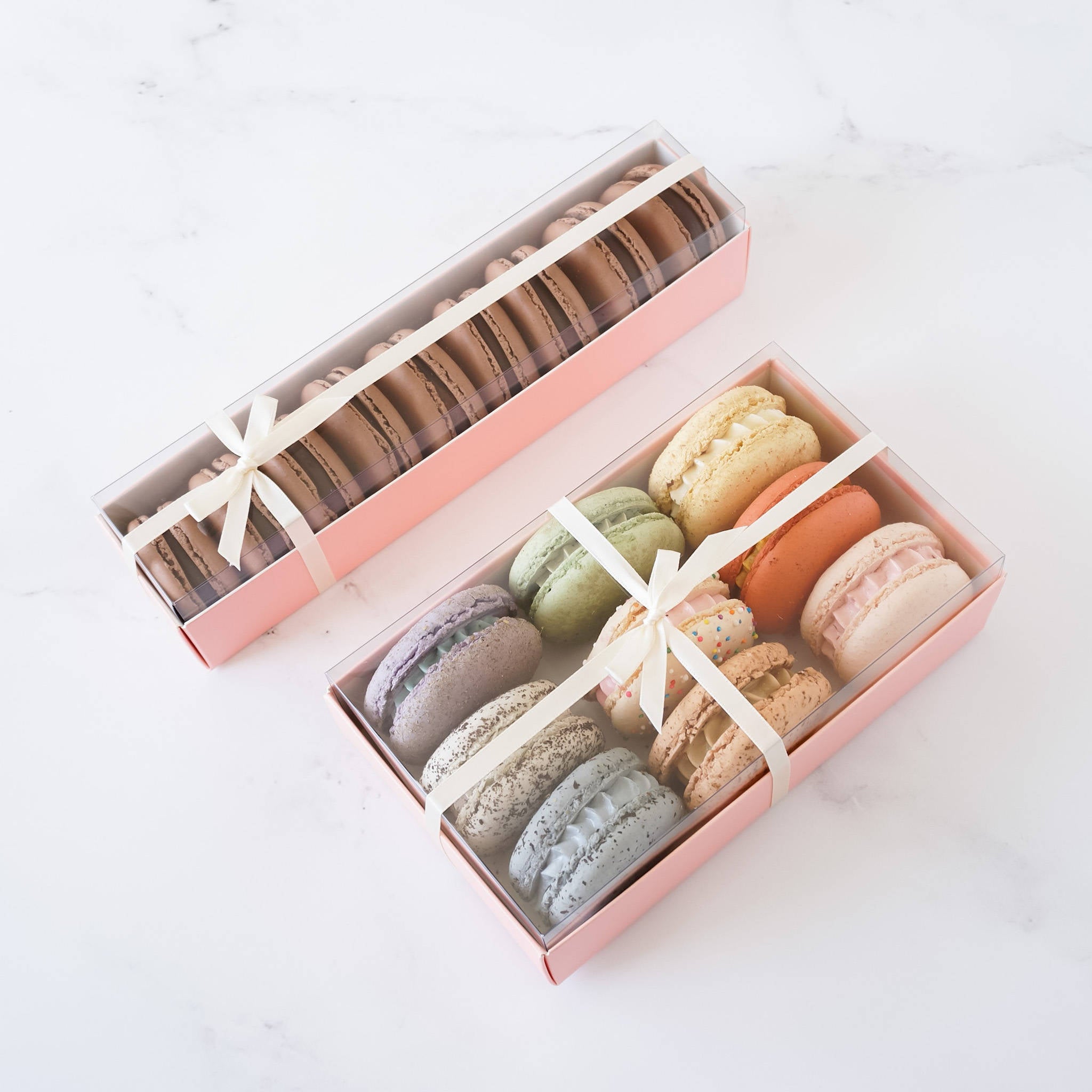 Two pink boxes filled with macarons. The larger box contains 9 brown macarons and the smaller box contains 9 colorful macarons.