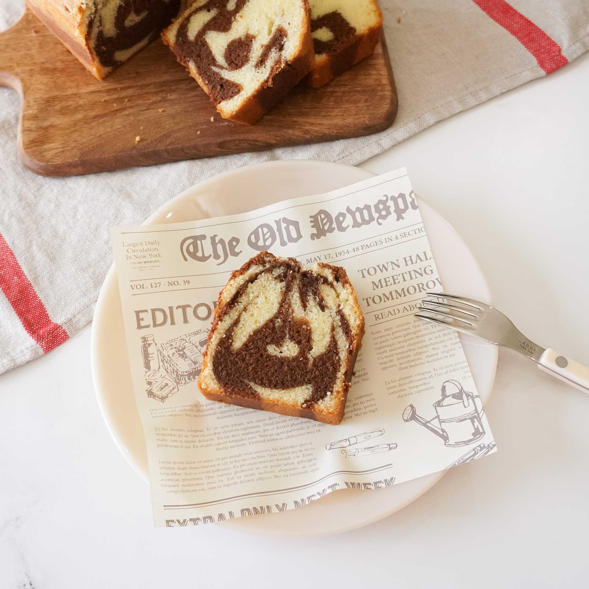 A piece of deli paper with a vintage newspaper theme is placed under a slice of marble cake on a plate, with a fork beside it.