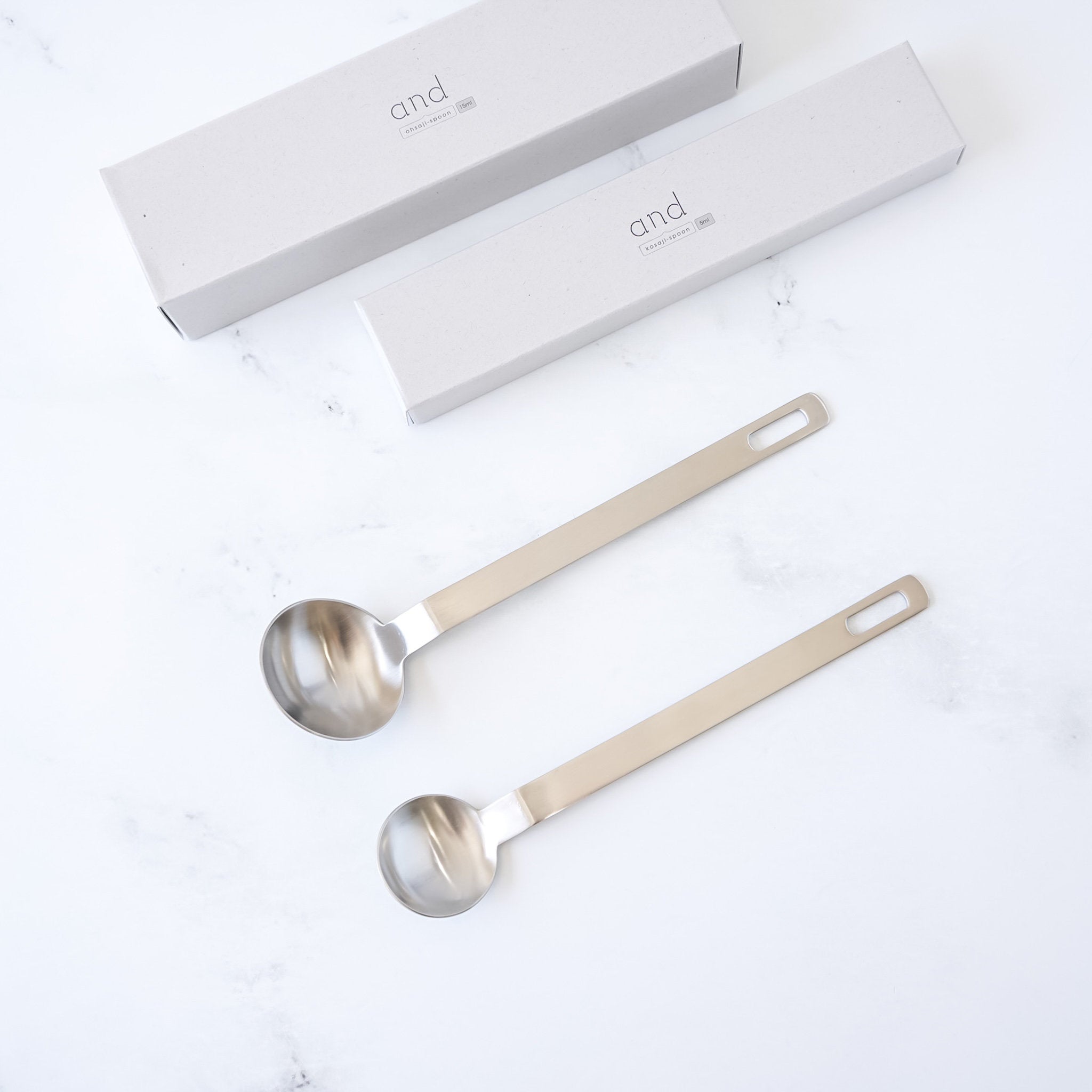 Two stainless steel measuring spoons with long handles on a white background, accompanied by a branded box.