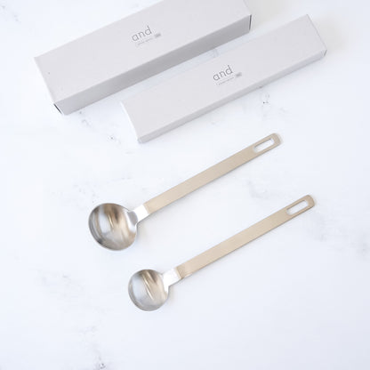 Two stainless steel measuring spoons with long handles on a white background, accompanied by a branded box.