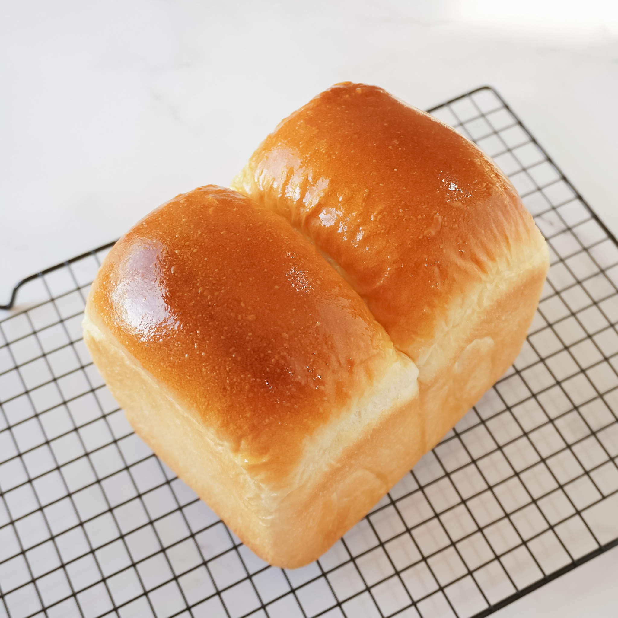 A loaf of freshly baked bread on a wire rack. The bread is golden brown and has a shiny, glazed surface. The bread is divided into two sections, with a seam running down the center.