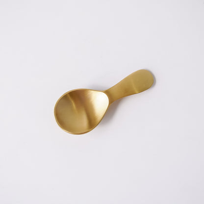 A small gold-colored metal spoon with a flat, matte finish, photographed diagonally on a clean white background.
