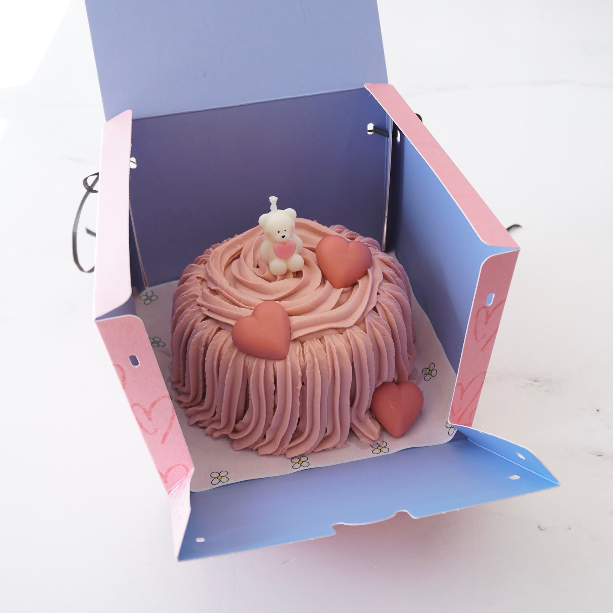 A pink box with a heart pattern and black ribbon handles. The box is open and contains a pink cake with a white teddy bear candle on top. The cake is decorated with pink frosting and three heart-shaped chocolates.
