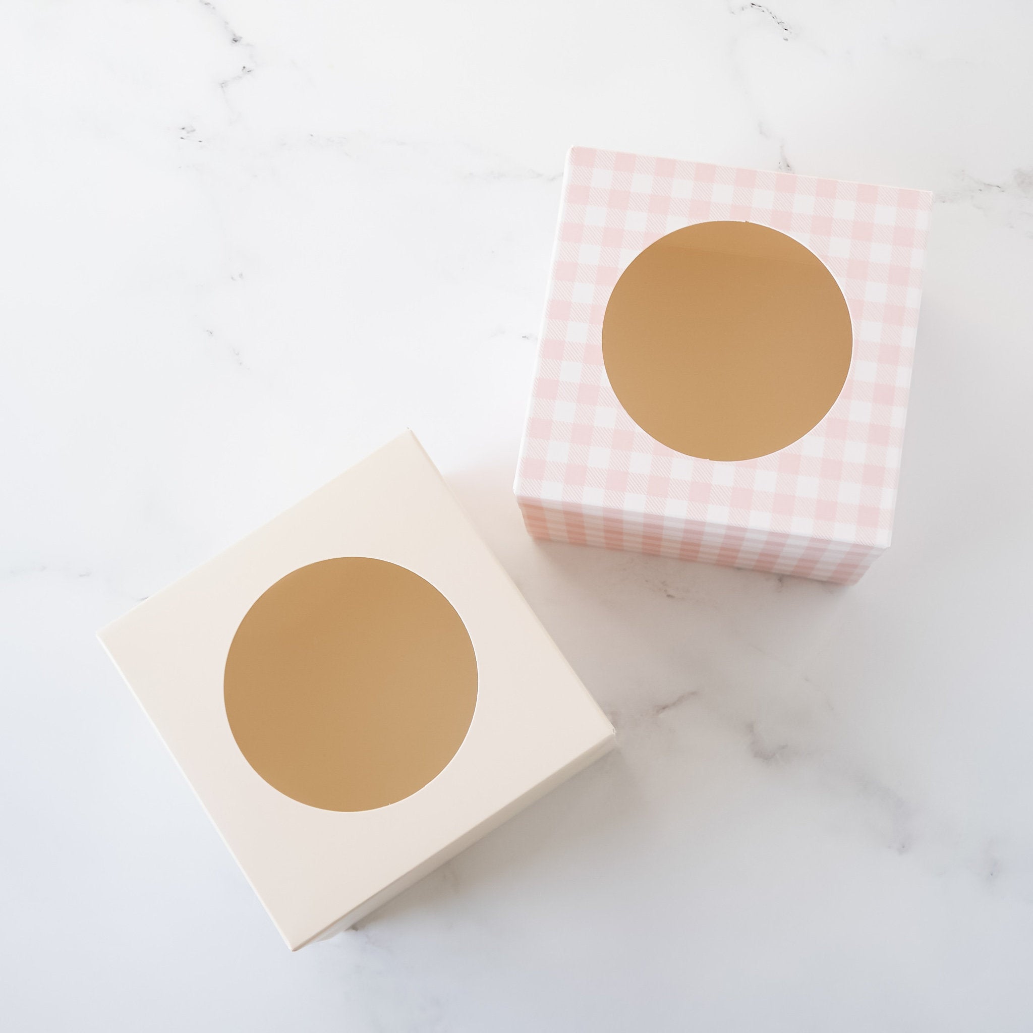 Two square boxes with round cutouts. One box is ivory and the other is pink with a white gingham pattern.